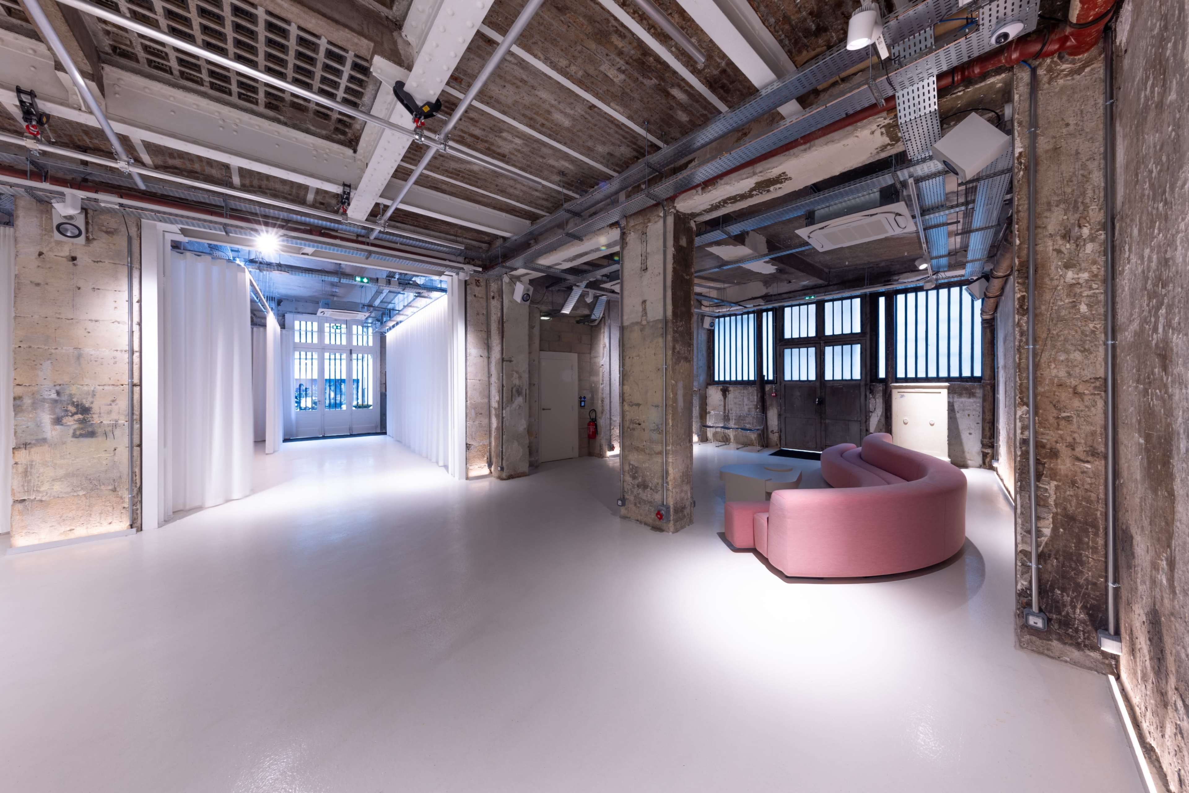 The image shows a modern industrial-style interior space with a pink sofa, open layout, and exposed building elements such as concrete walls and overhead beams.