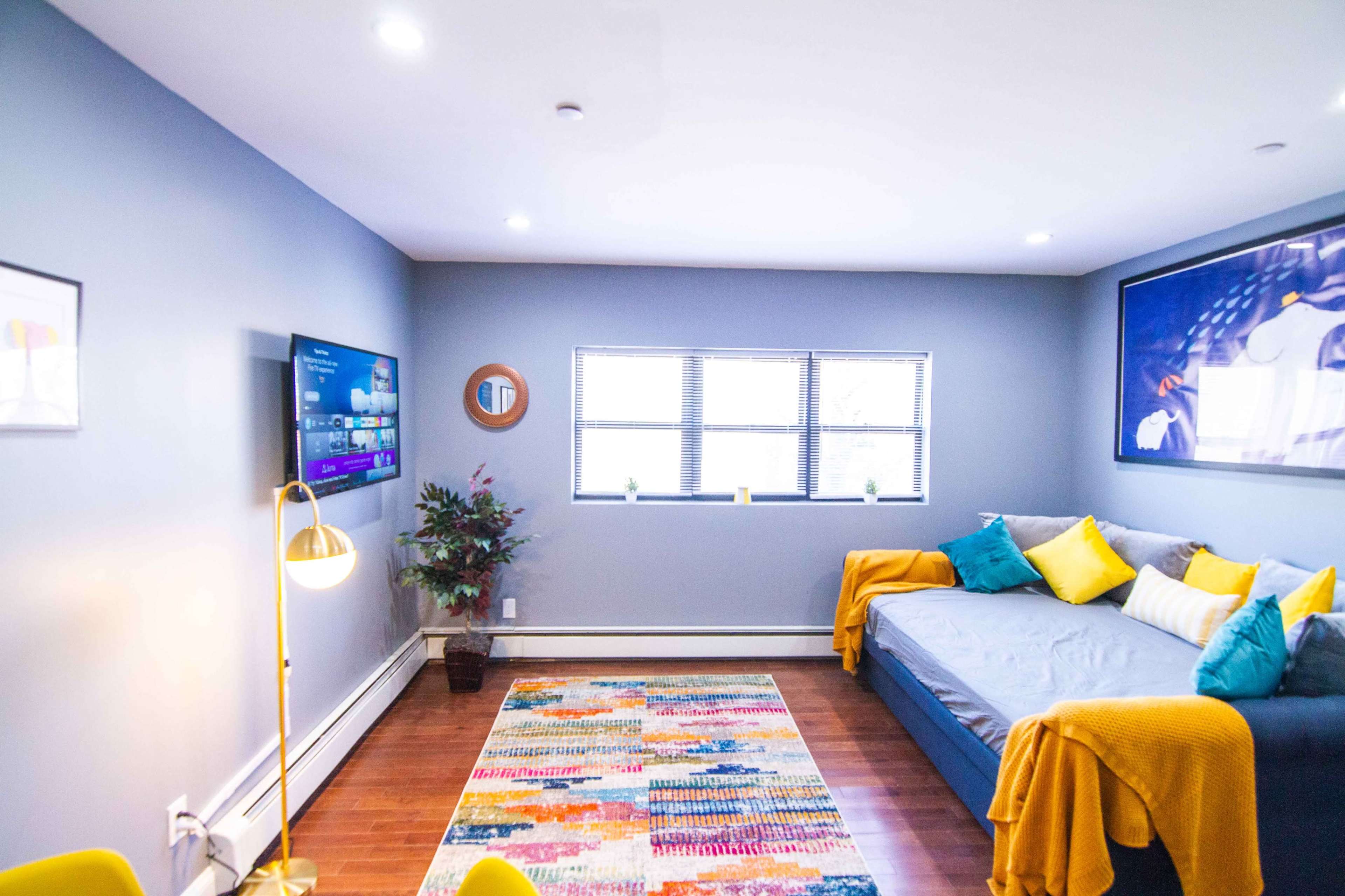 A modern living room featuring a gray wall, a television mounted on one wall, a daybed with colorful pillows, and a patterned area rug.