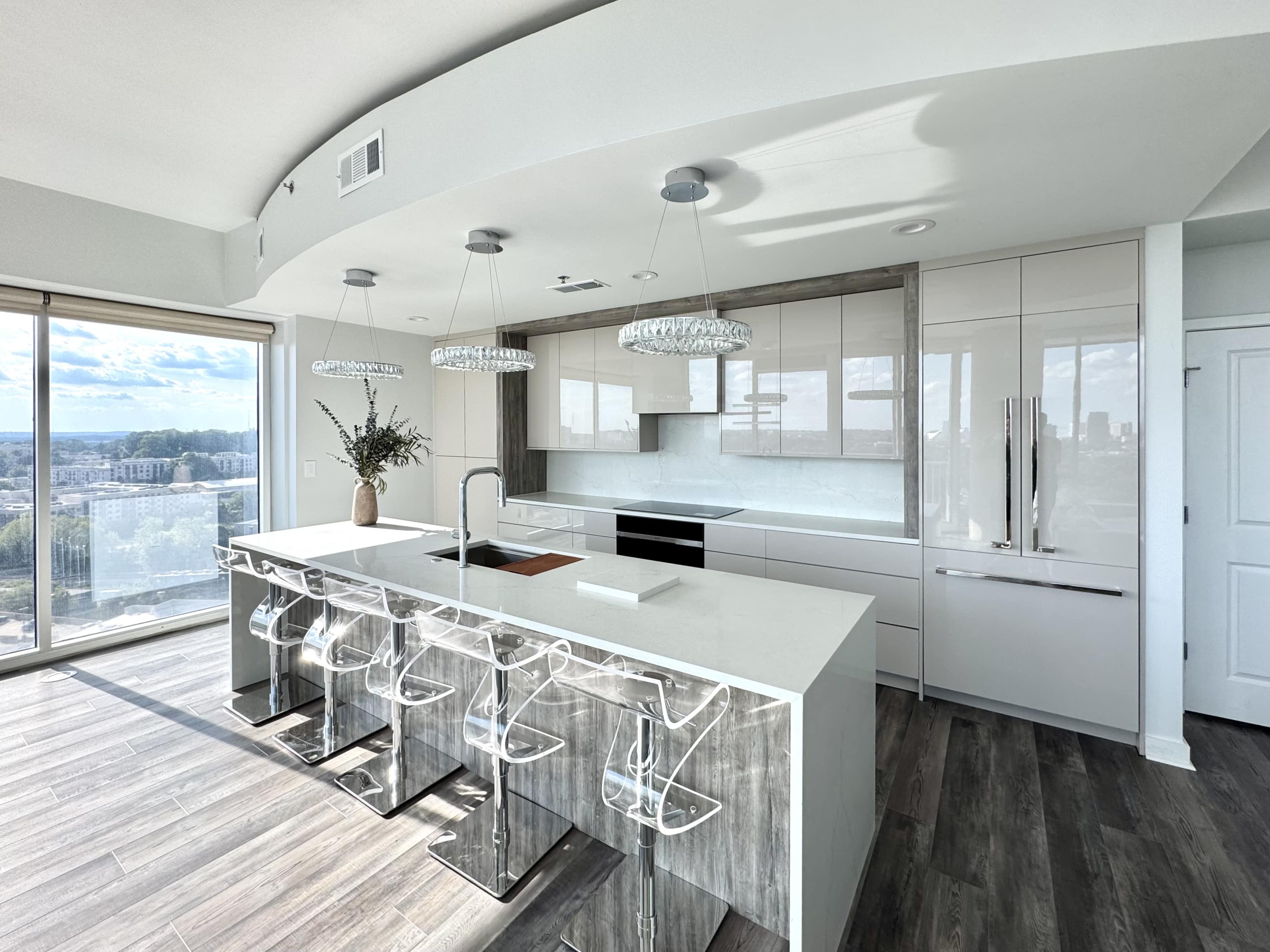 The image shows a modern kitchen with a large island, sleek cabinetry, and several bar stools, overlooking a city skyline through large windows.