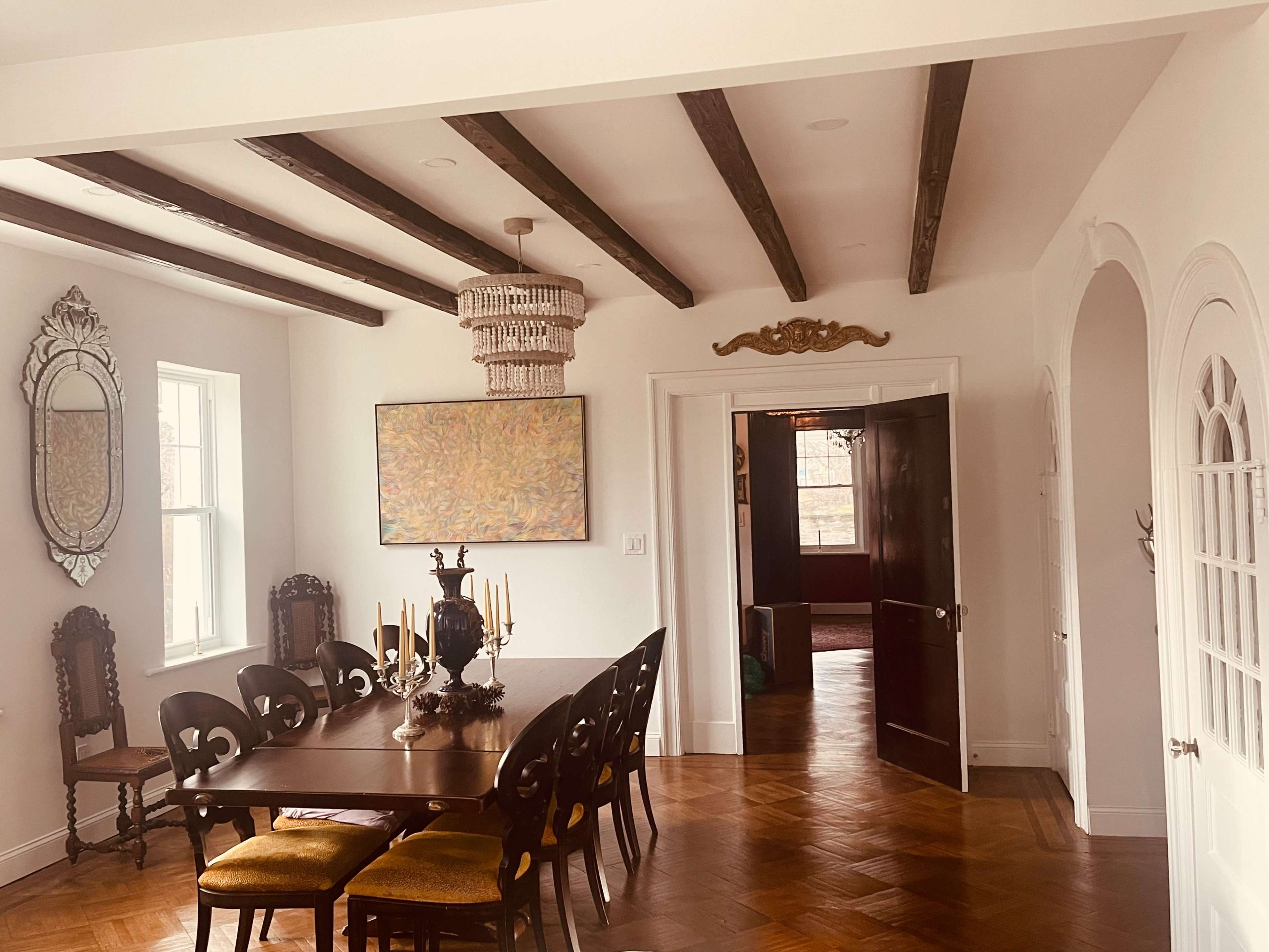 A dining room features a long wooden table surrounded by chairs, with a chandelier above and an ornate mirror on the wall.