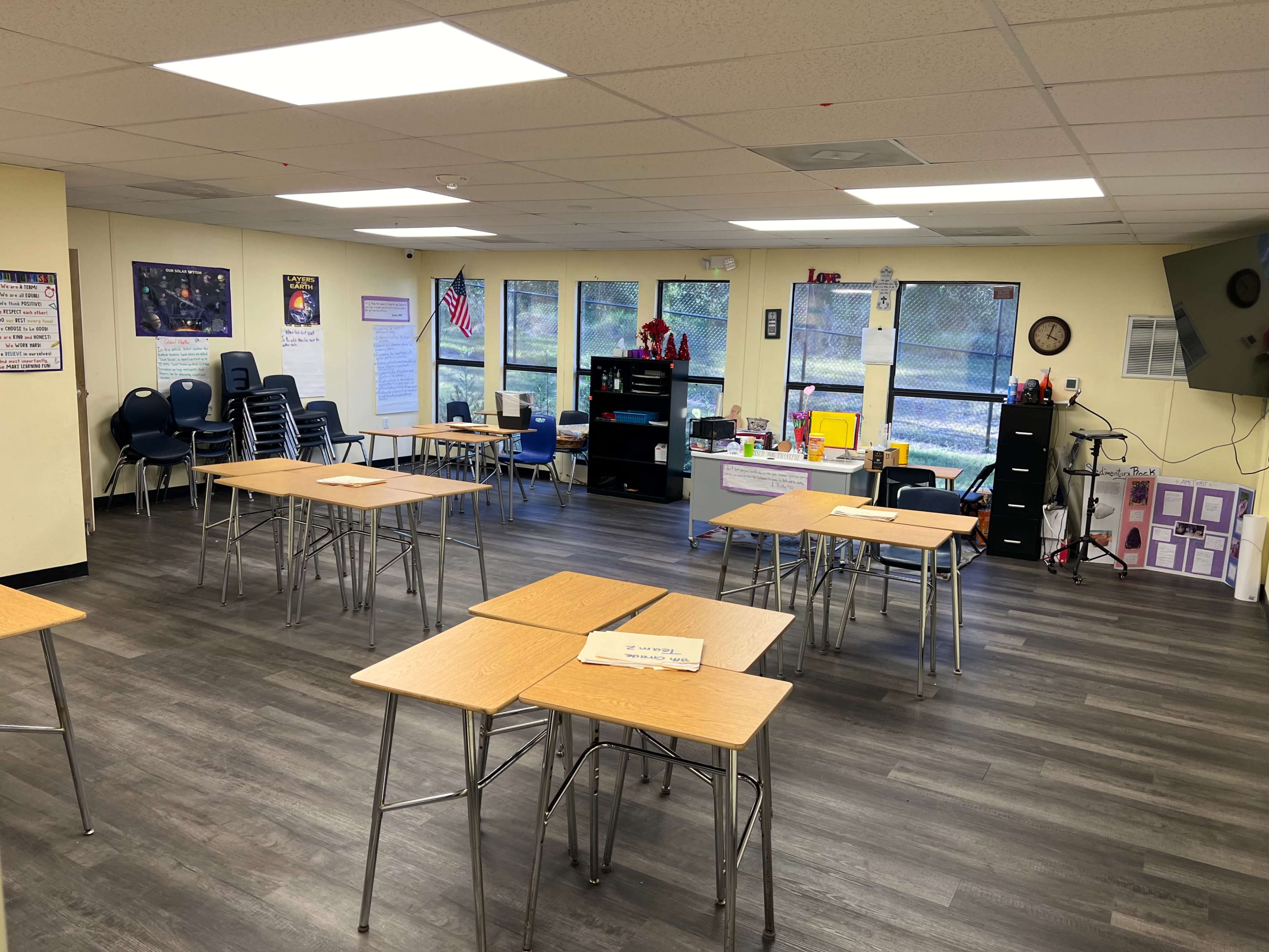 A classroom with several empty tables arranged in rows and various educational resources displayed on shelves and walls.