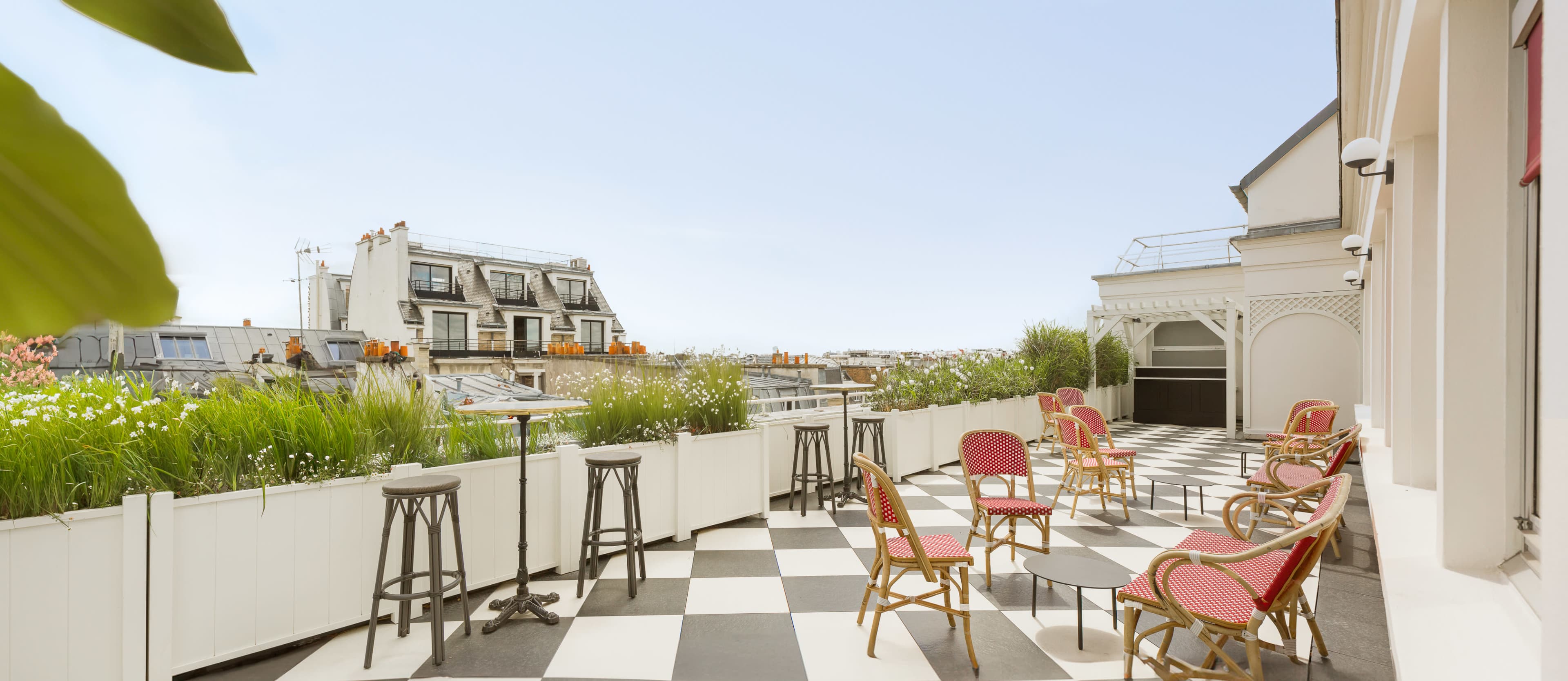 A rooftop terrace featuring checkerboard flooring, tables, and chairs with a view of surrounding buildings and greenery.