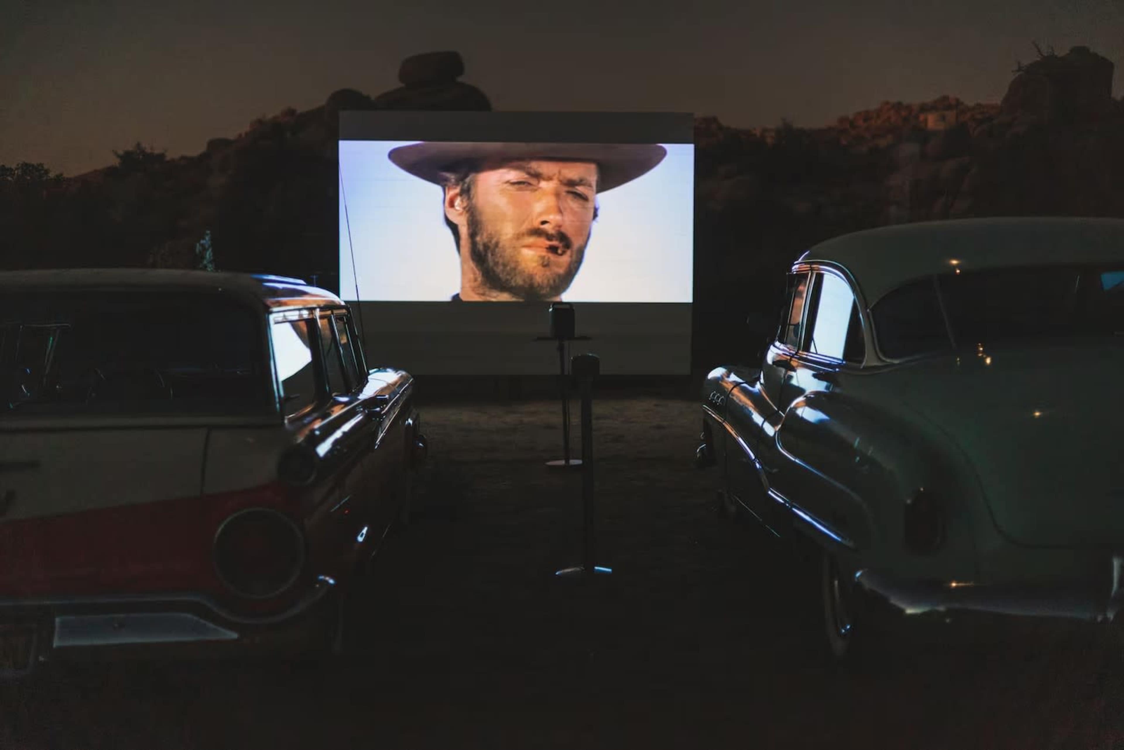 The image shows two classic cars parked in front of a large movie screen displaying a scene featuring a man wearing a hat.