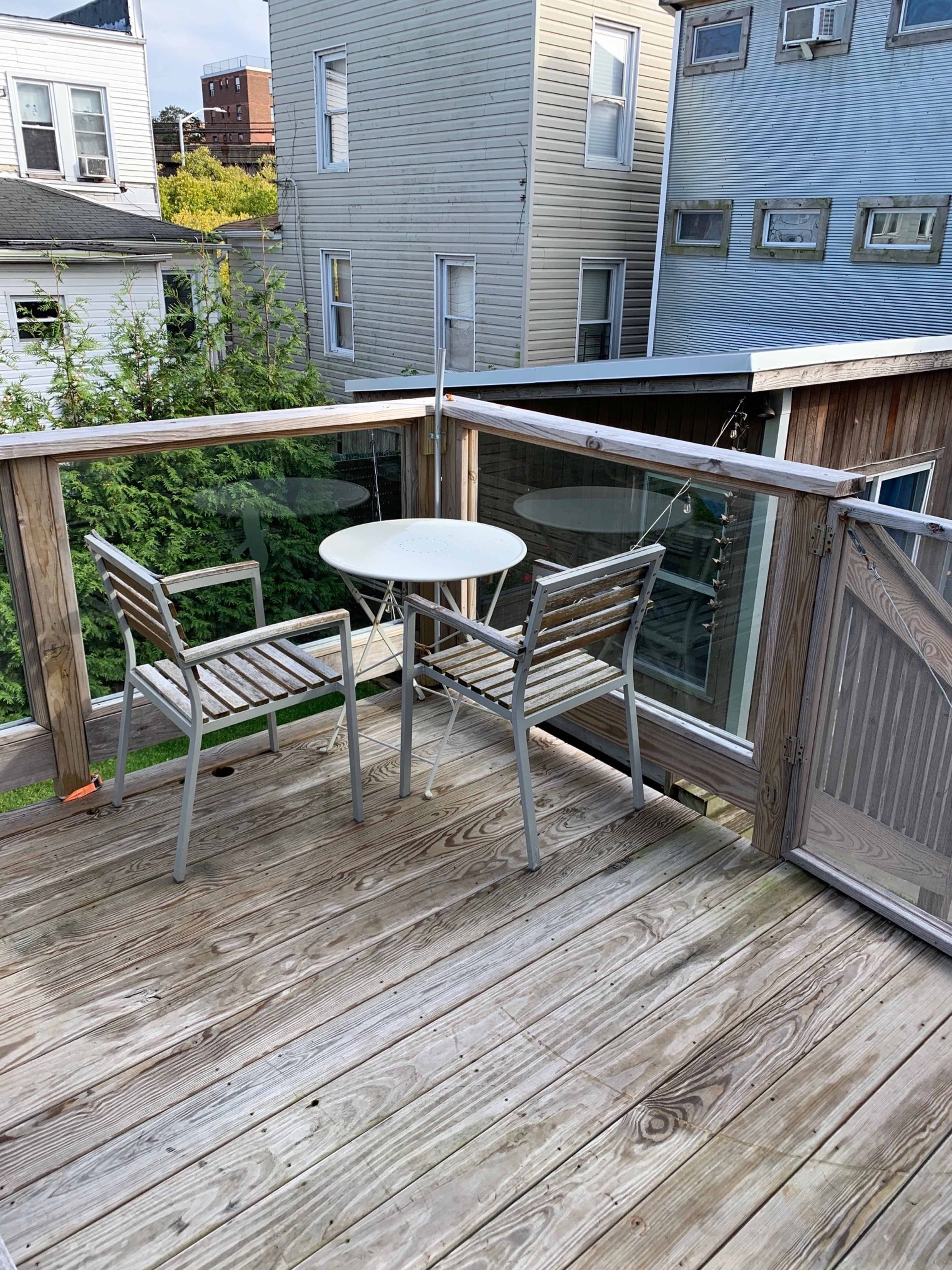 The image shows a small balcony with a round table and two wooden chairs, surrounded by wooden railings and facing neighboring buildings.