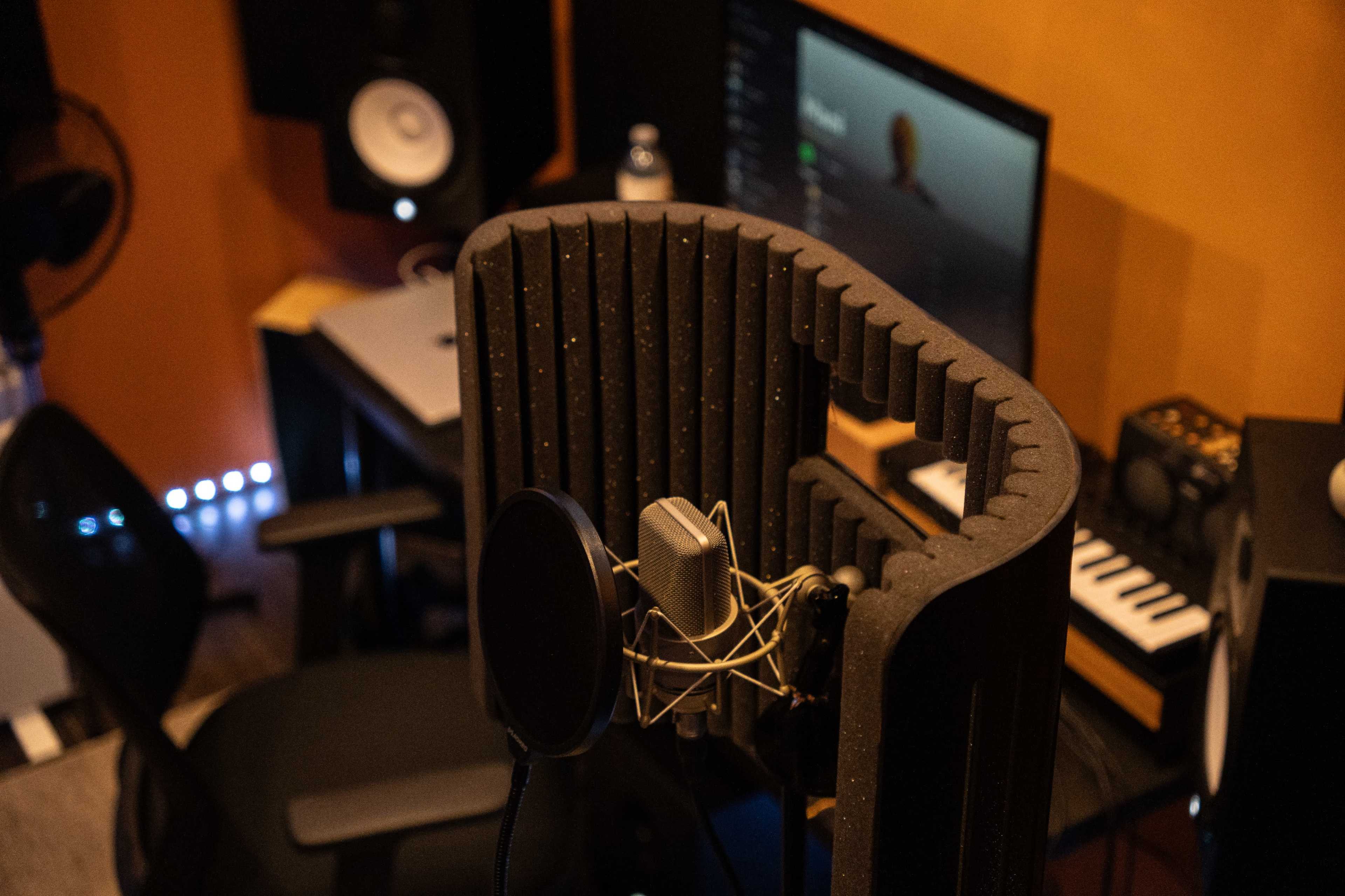 A microphone is positioned in a soundproofed area with a computer and audio equipment visible in the background.