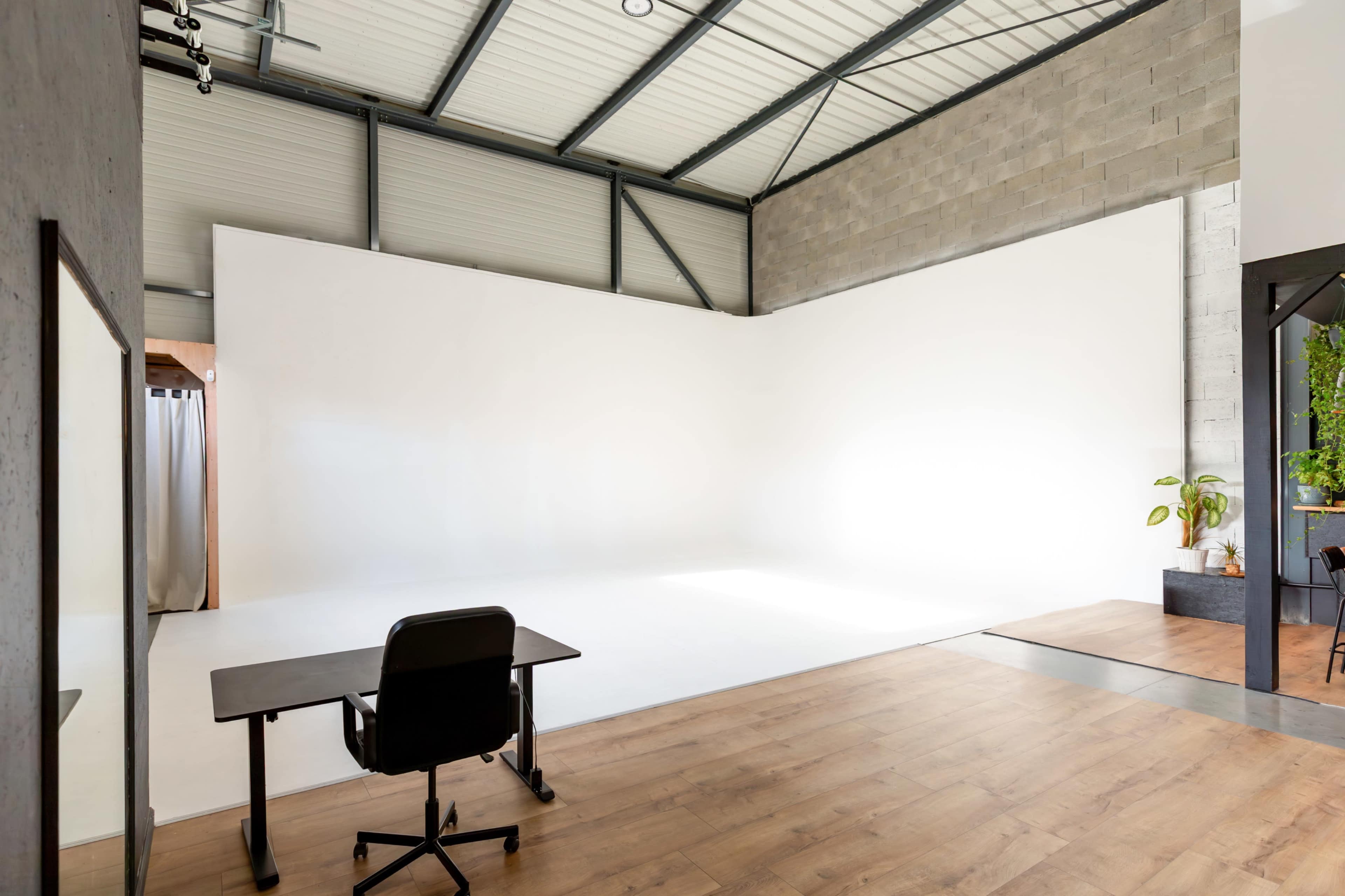 The image shows a spacious, well-lit studio with a large white wall, a wooden floor, and a black office chair at a desk.