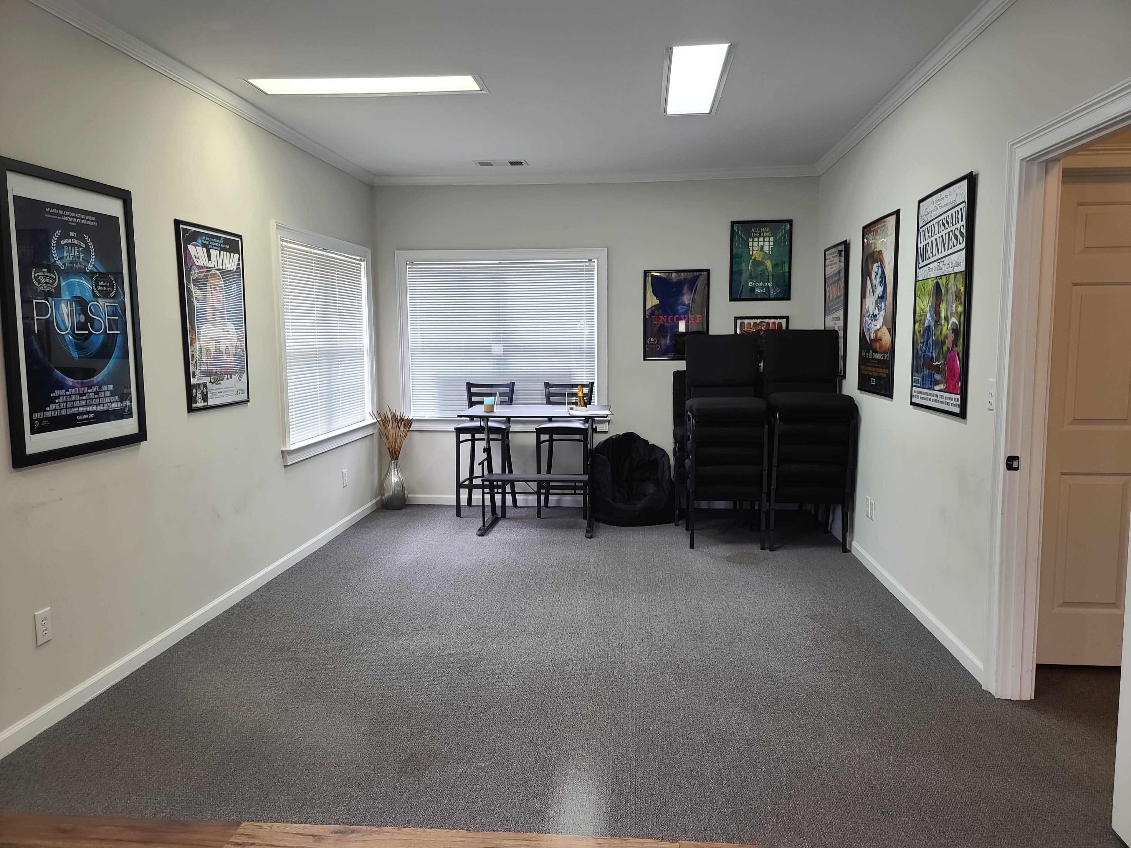 A spacious room with gray carpet, featuring a table with chairs, stacked chairs against the wall, and several framed posters on the walls.