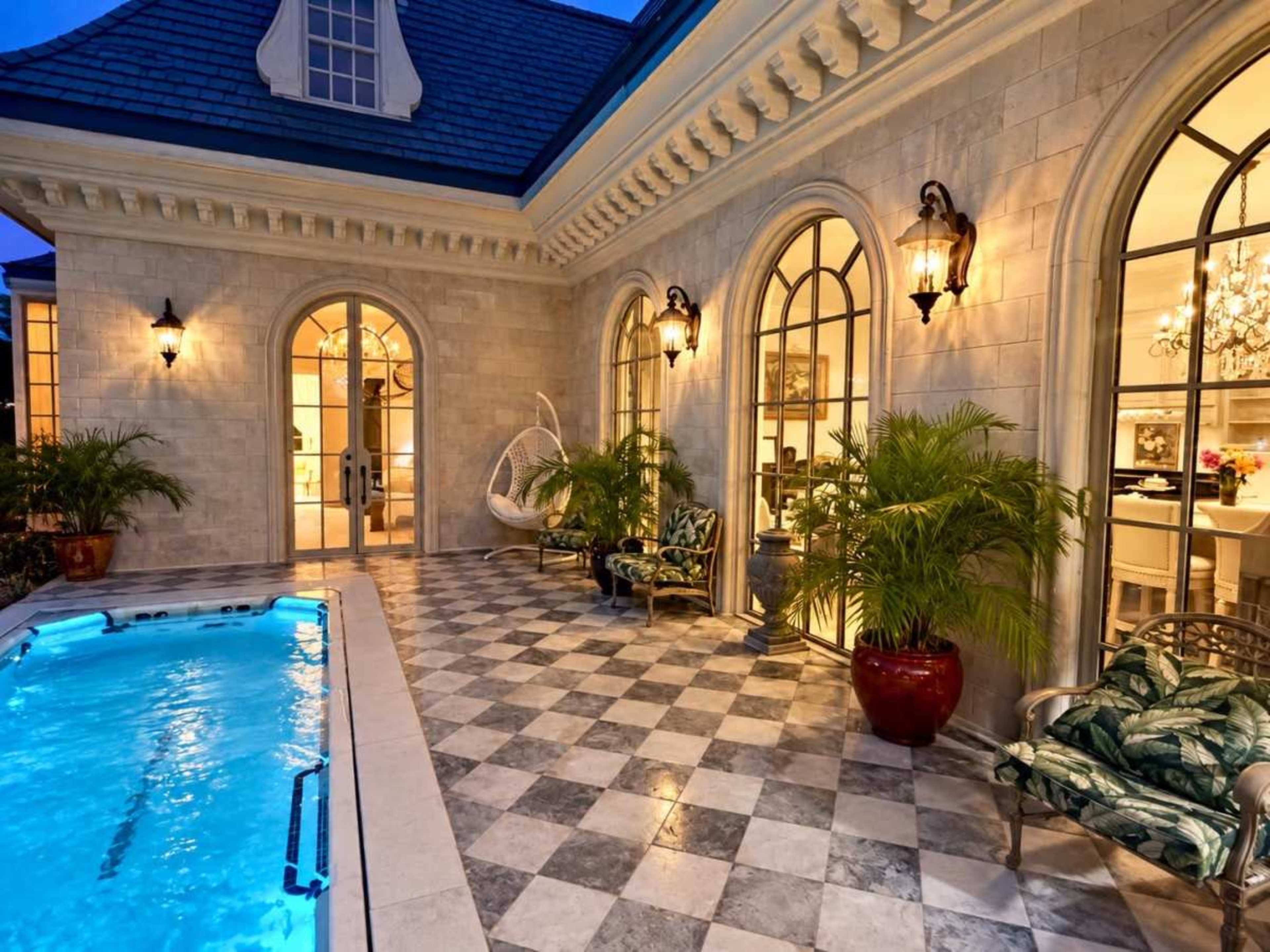 The image shows a stylish outdoor patio area featuring a small pool, checkerboard tile flooring, and elegant furniture surrounded by large windows and tropical plants.