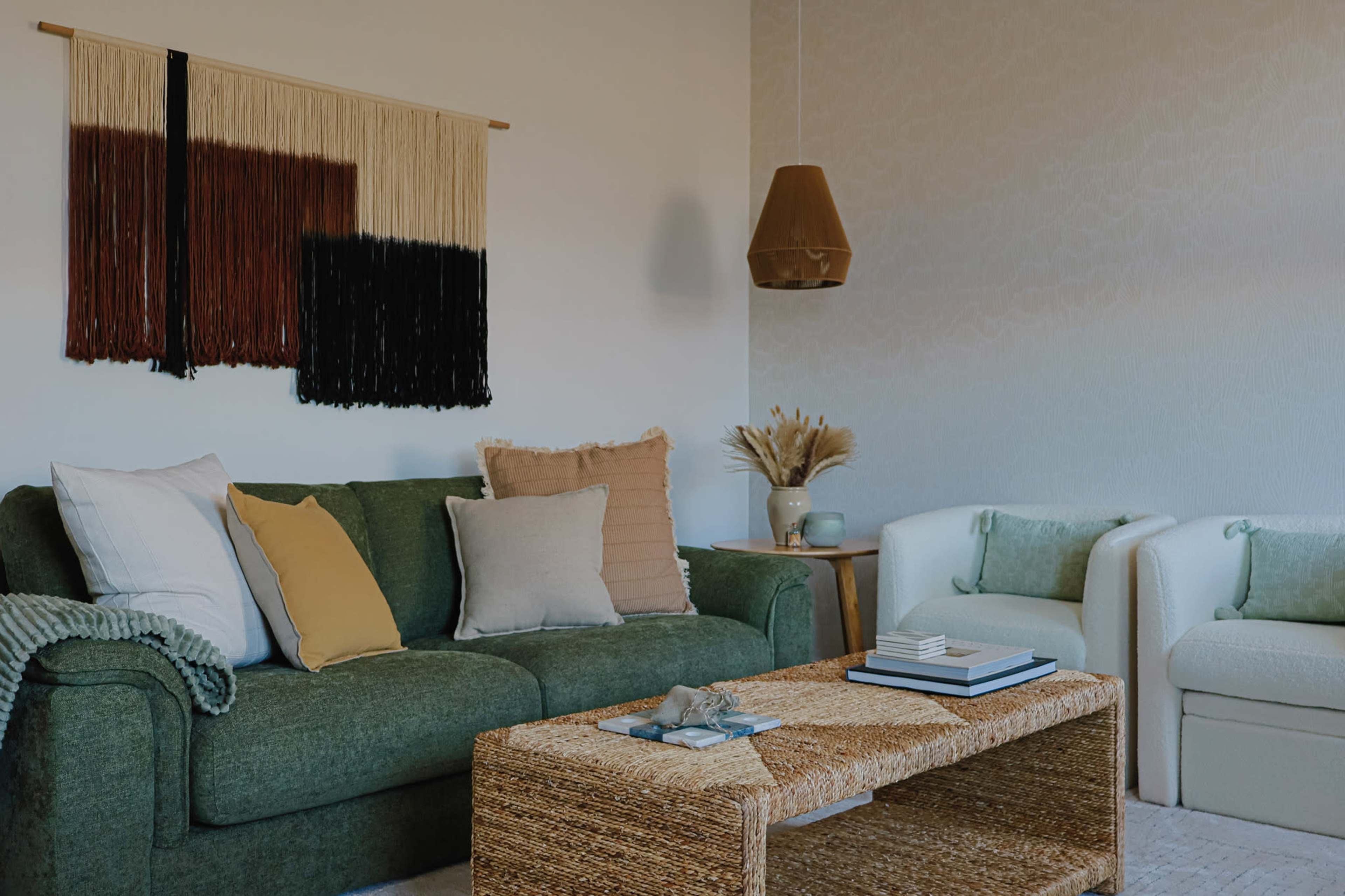 The image shows a cozy living room with a green sofa adorned with various pillows, a woven coffee table, and a light-colored armchair, accompanied by a decorative wall hanging and a pendant light.
