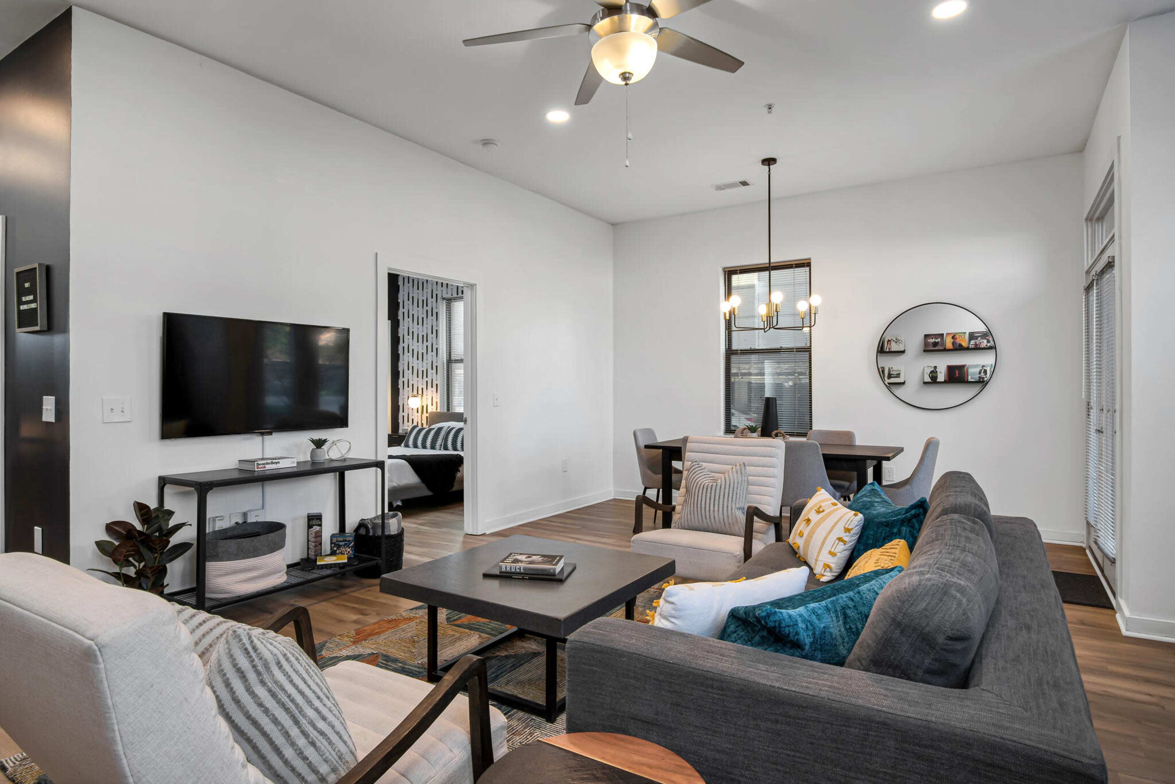 The image shows a modern living room with a sectional sofa, a coffee table, a television on a stand, and a dining area with a round table and chairs in the background.