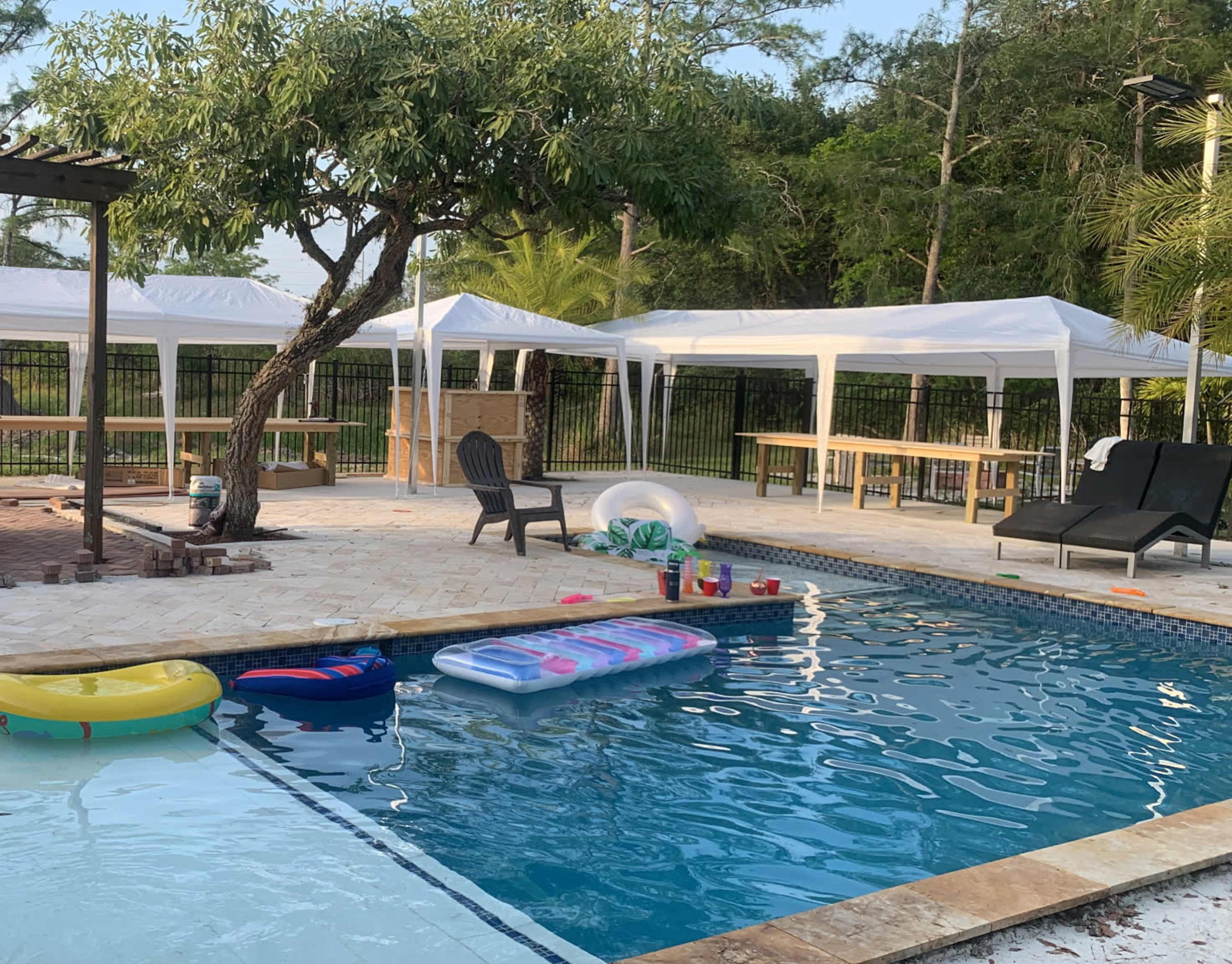 The image shows a backyard pool area with floating pool toys, lounge chairs, and white canopies set up nearby.