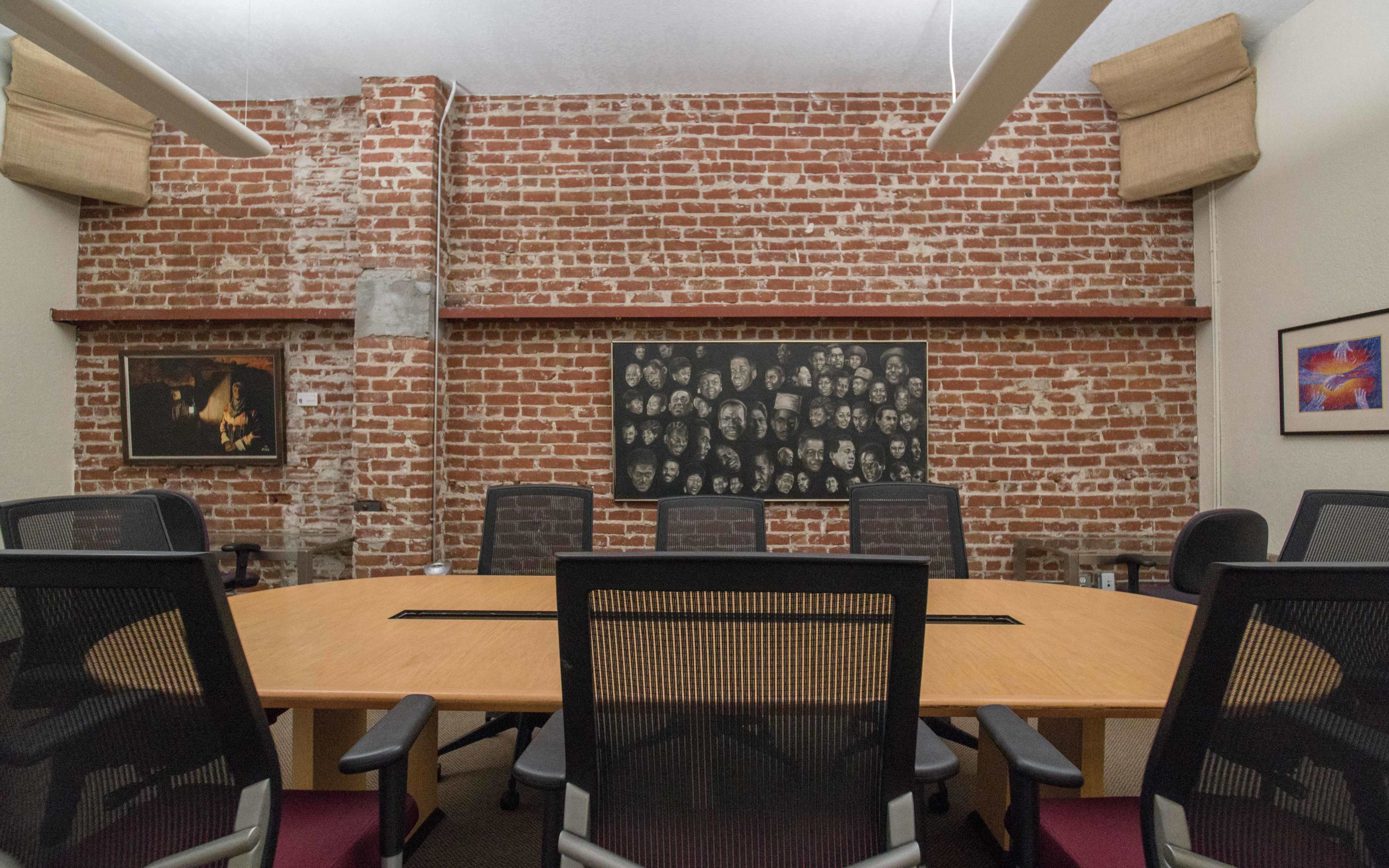 A conference room features a large wooden table surrounded by black chairs, with a red brick wall decorated with framed art and a collage of faces.