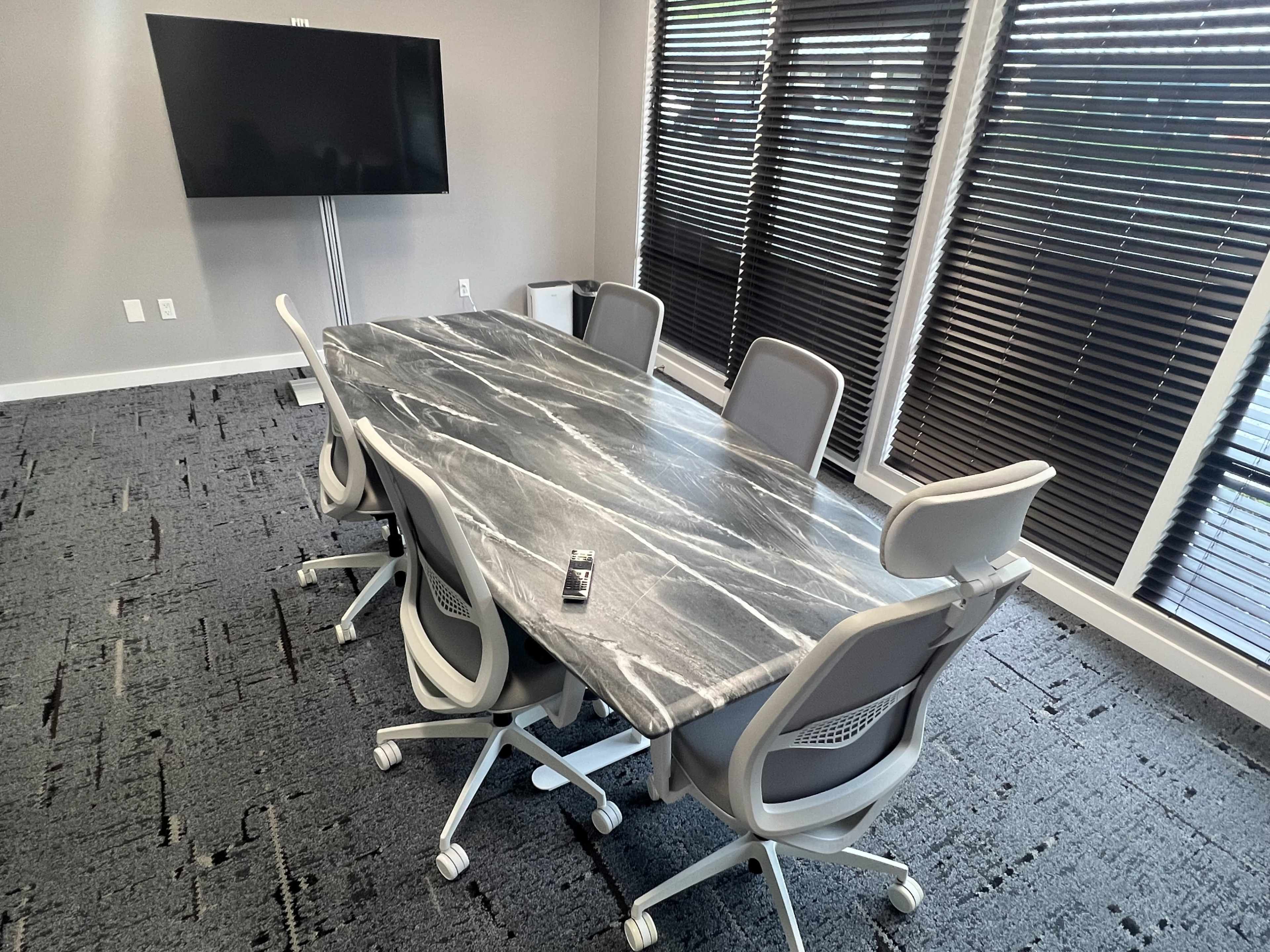 A marble conference table surrounded by five ergonomic chairs is situated in a brightly lit meeting room with large windows covered by blinds.
