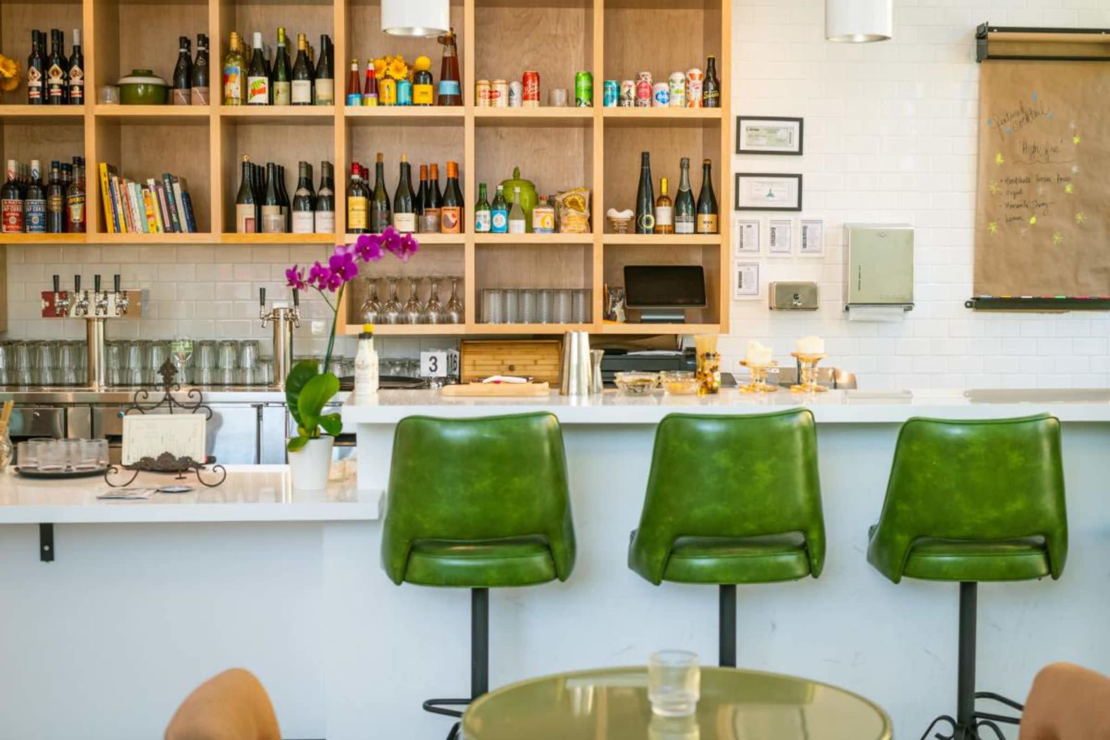The image shows a modern bar area with green bar stools, a white countertop, and a wooden shelving unit displaying various bottles and canned beverages.