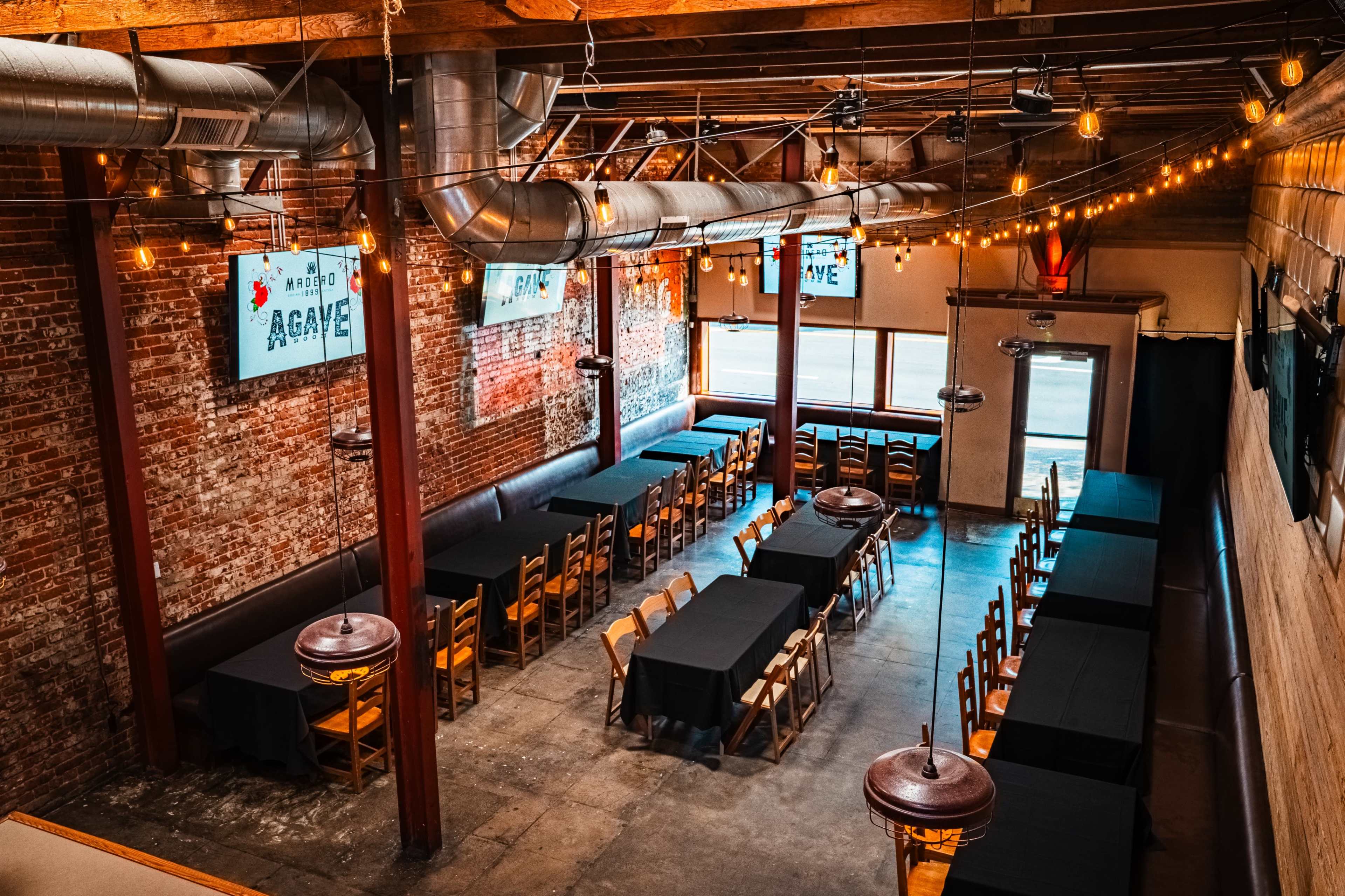 The image shows a spacious restaurant interior with wooden beams, exposed brick walls, and multiple long tables set with black tablecloths arranged under string lights.
