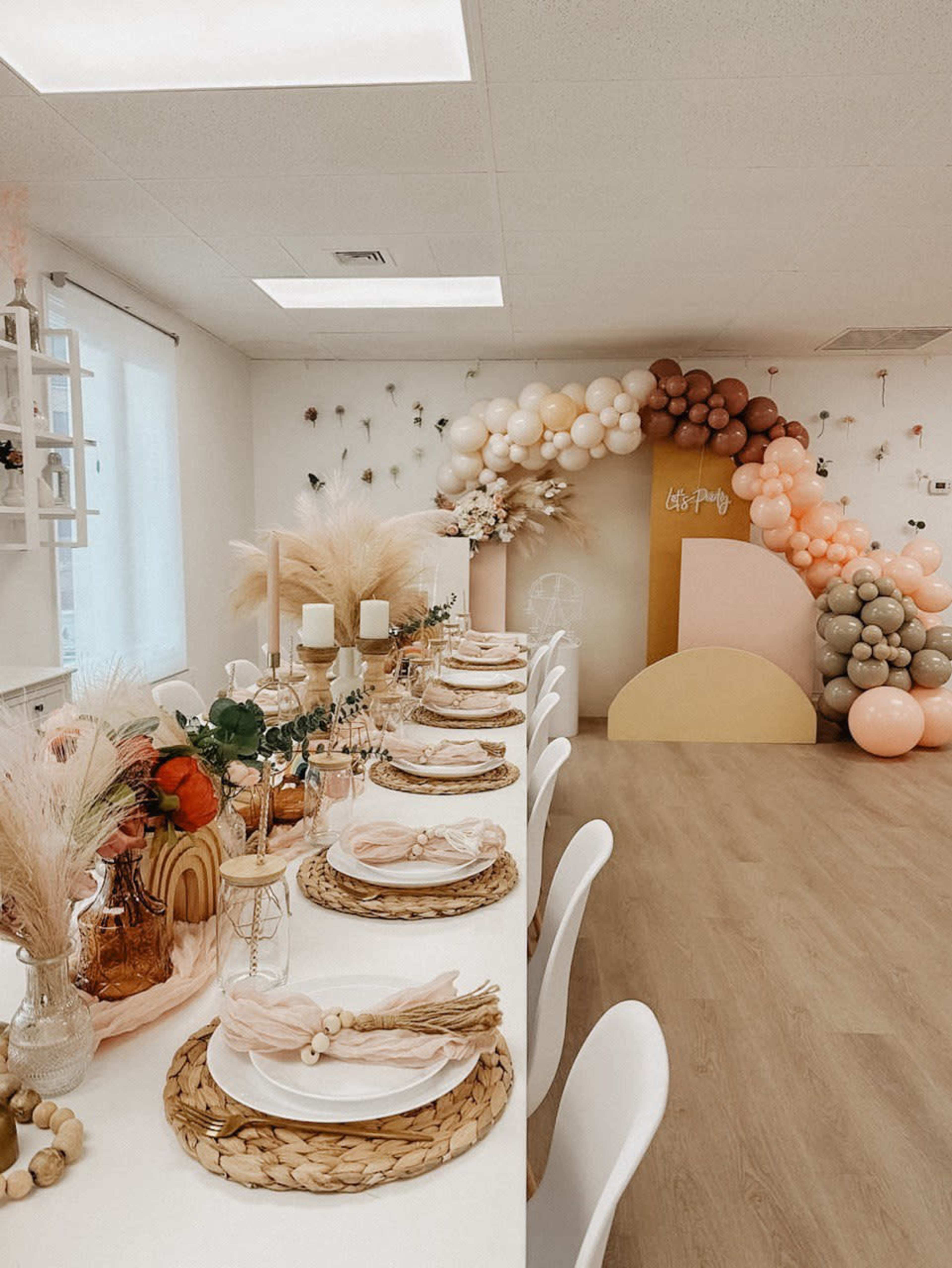 A decorated dining area featuring a long table set with pastel tableware, floral arrangements, and a colorful balloon arch in the background.