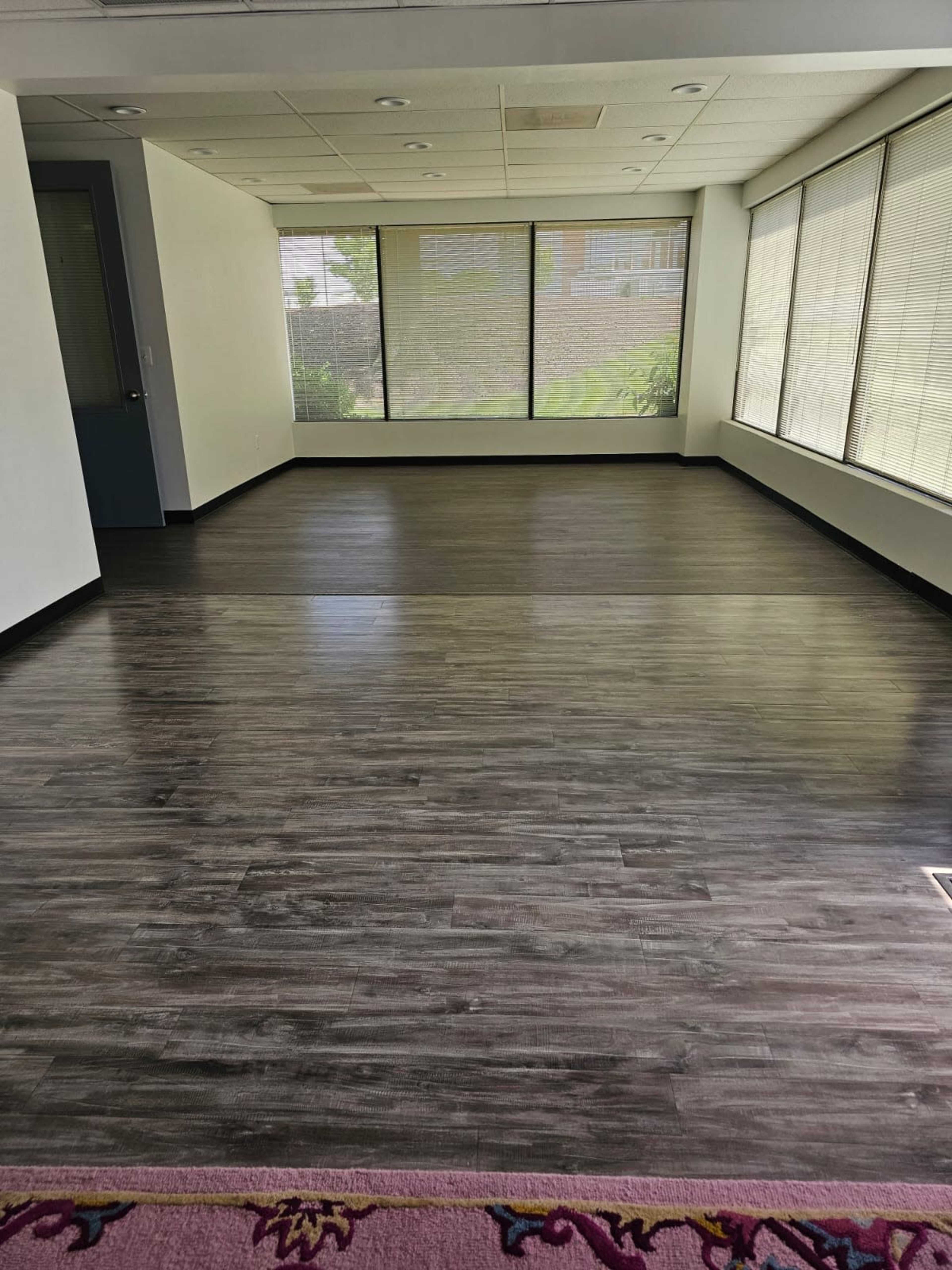 An empty room with dark laminate flooring and large windows covered with blinds.