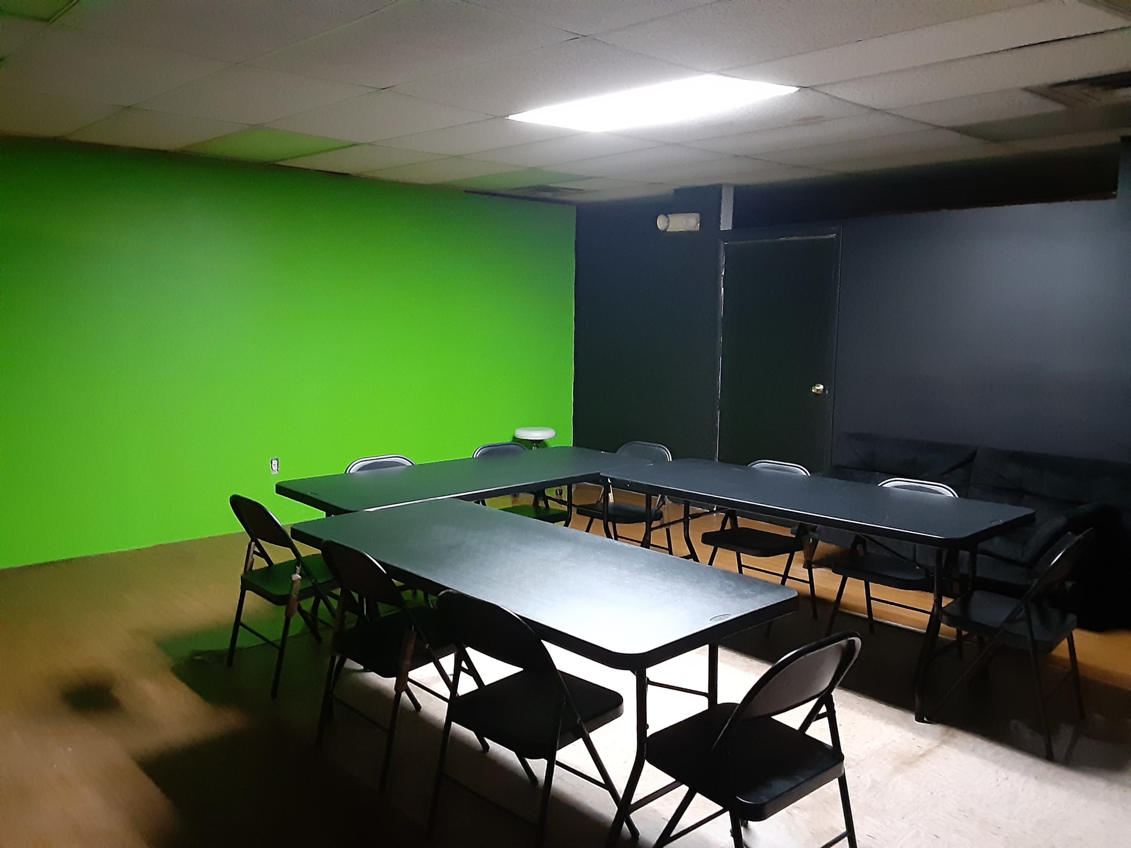 A dimly lit room with a green wall on one side, a black wall opposite, and a large table surrounded by chairs in the center.