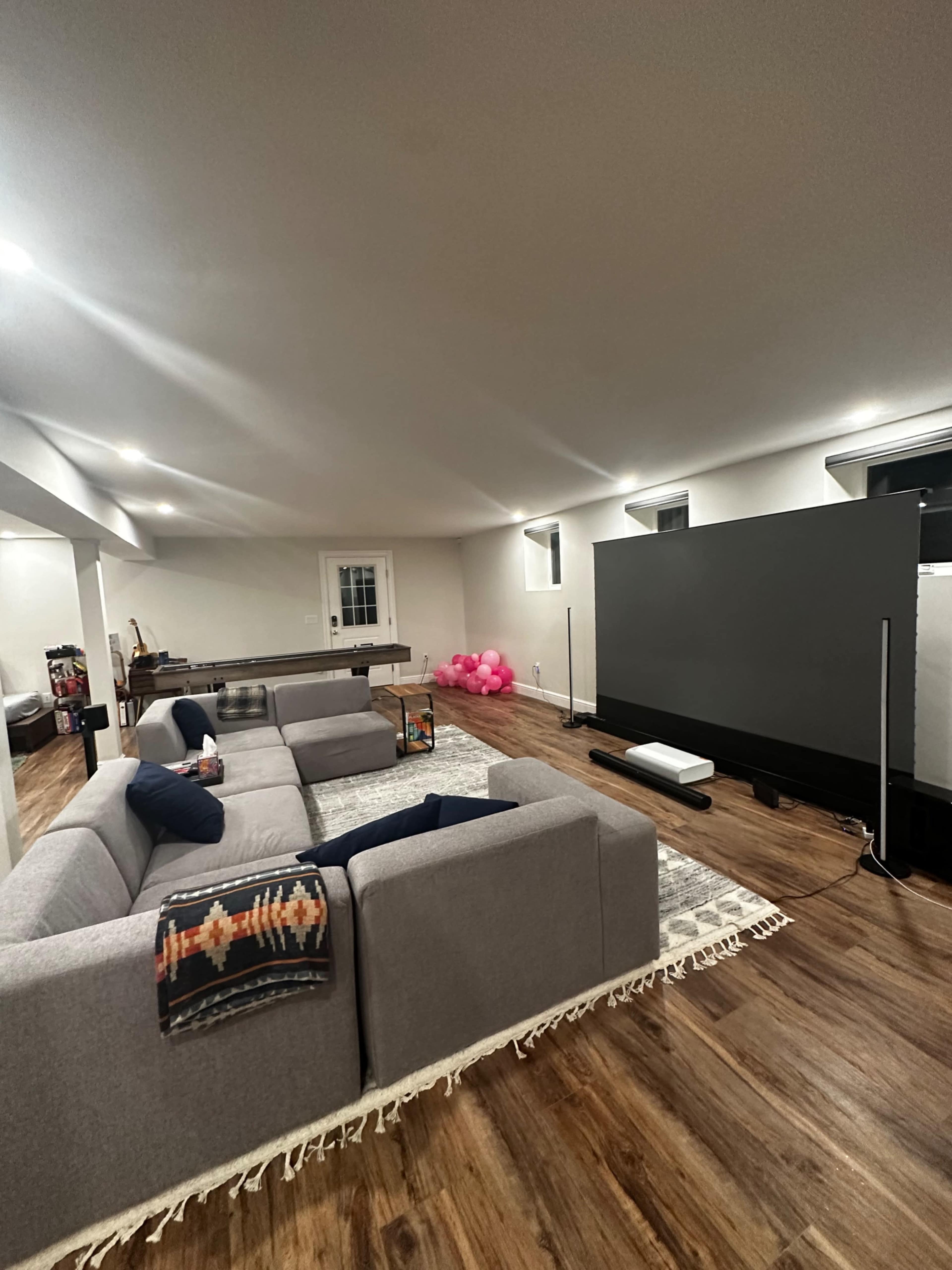 A spacious, well-lit basement living area featuring a large gray sectional sofa, a wooden coffee table, and a screen set up against one wall, with a cluster of pink balloons in the corner.