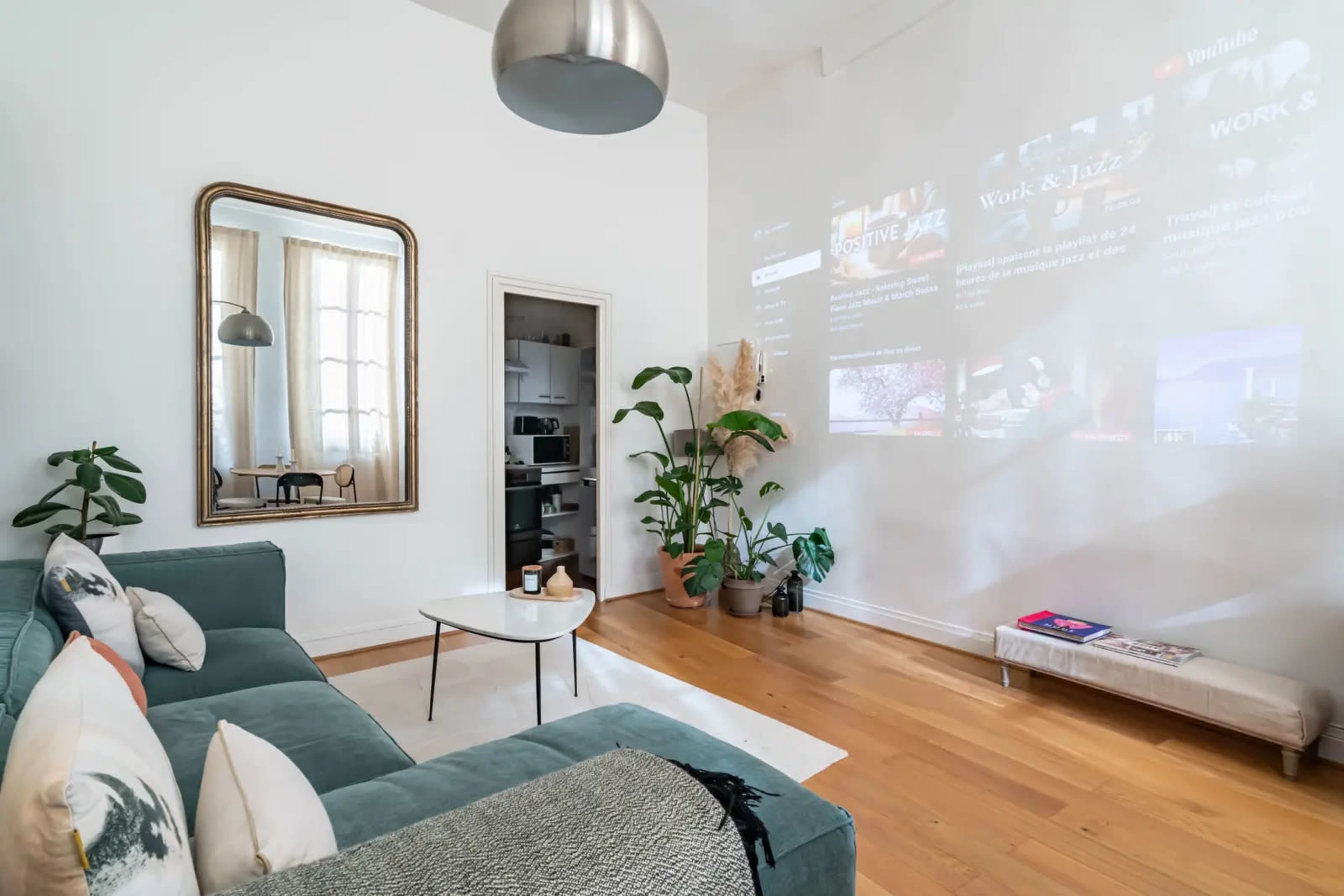 A bright living room features a green couch, a large mirror, and a projection on the wall displaying various media content.
