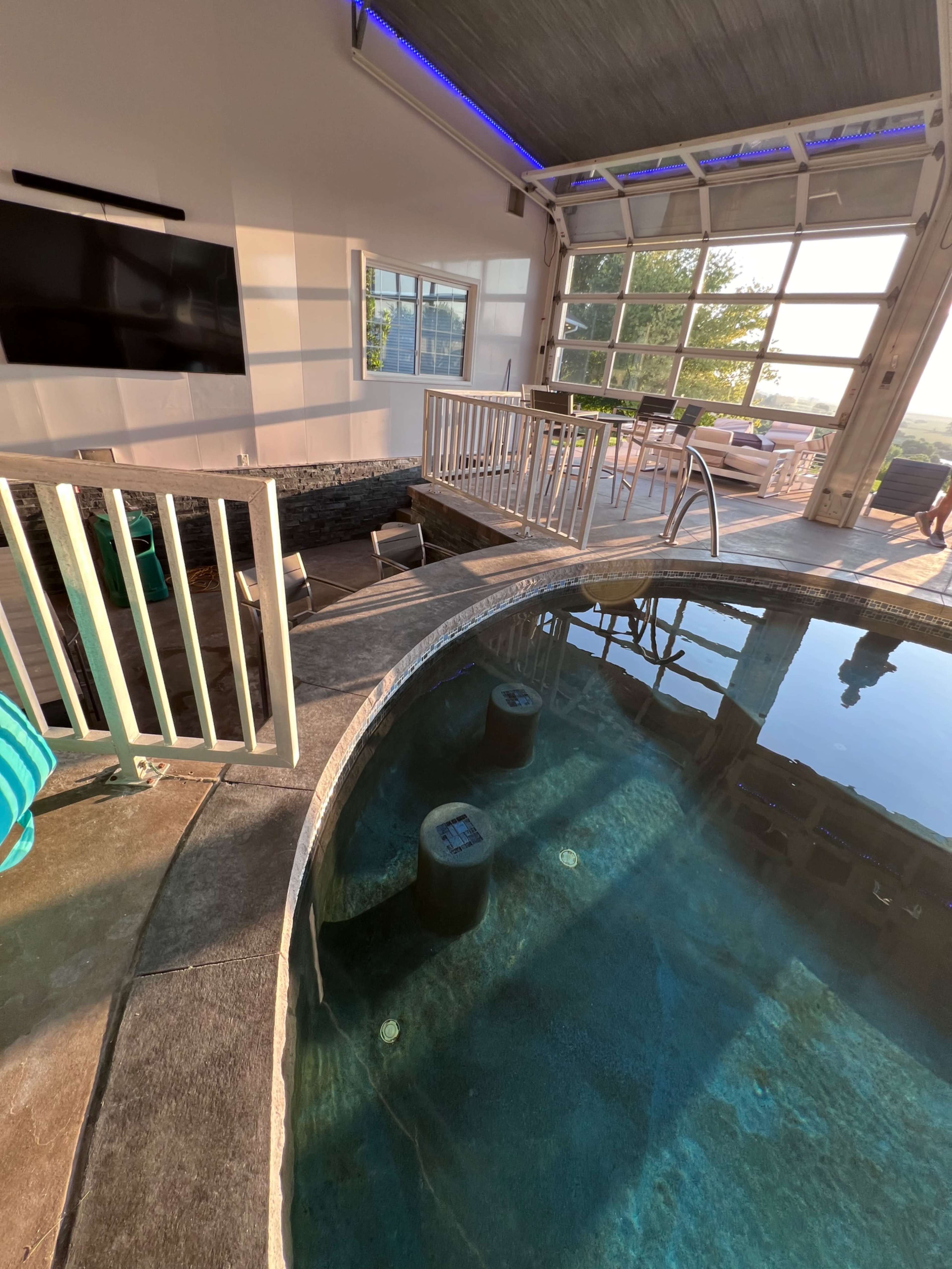 The image shows a modern indoor-outdoor space featuring a round hot tub, a flat-screen TV mounted on the wall, and a balcony with a view.
