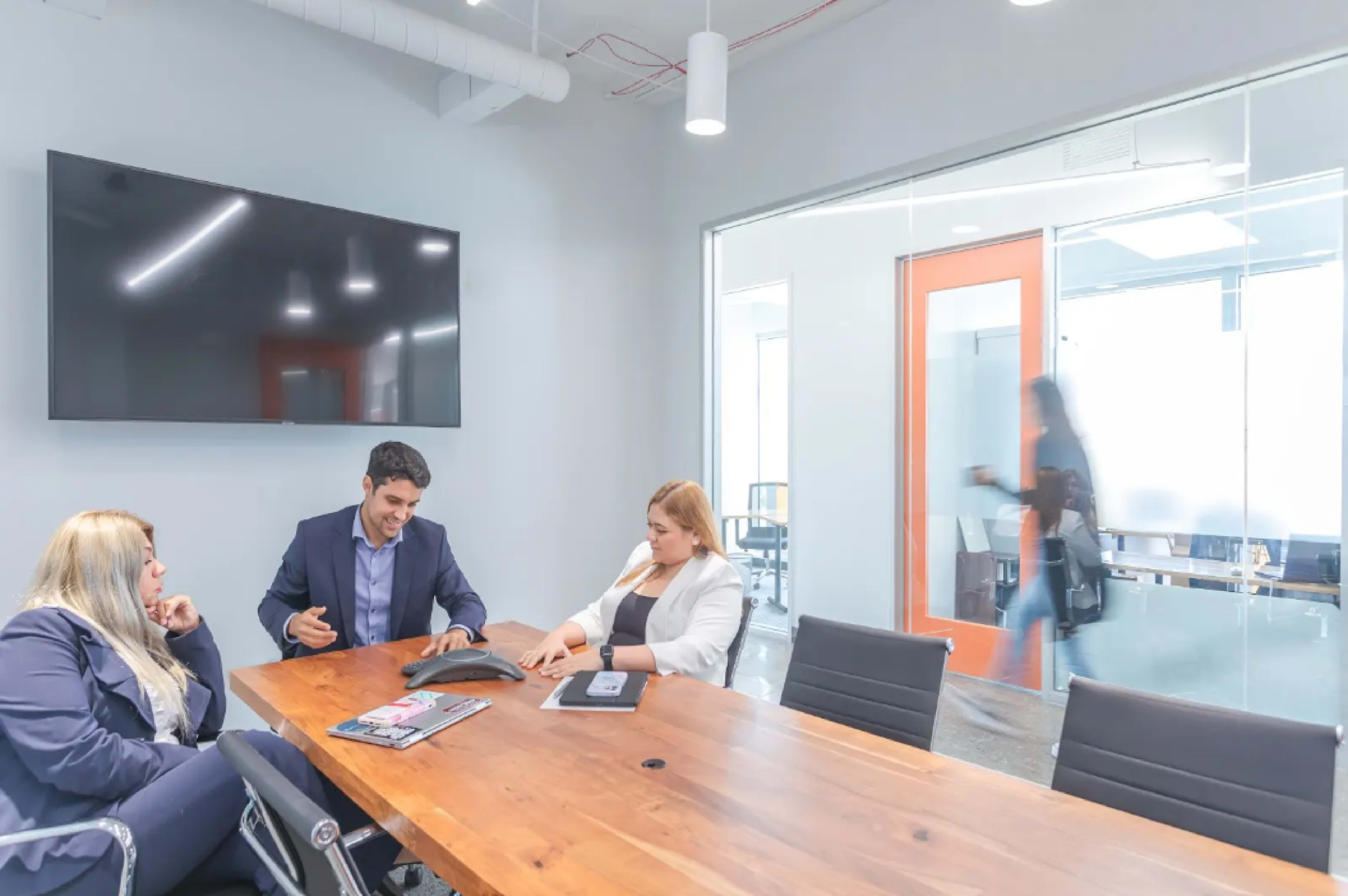 A group of three individuals is seated around a wooden conference table, engaged in discussion, while a person walks by in the background of a modern office setting.