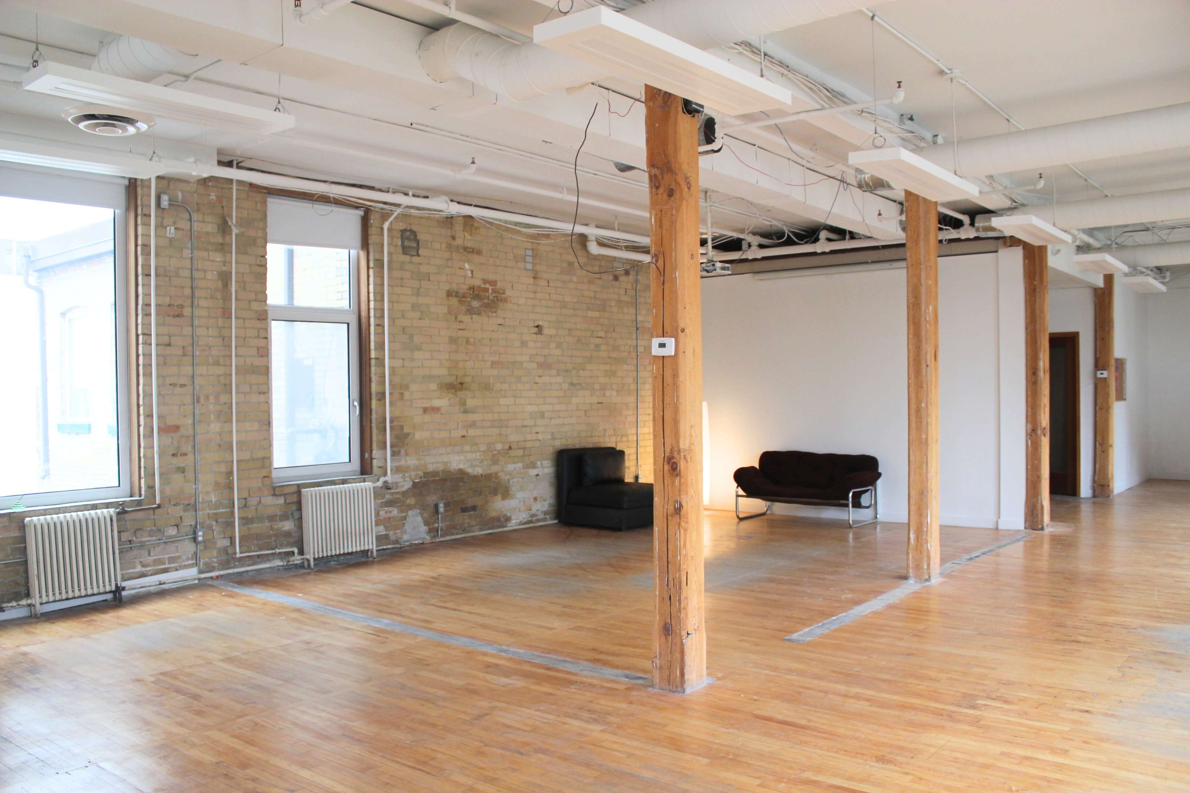The image shows a spacious, empty room with exposed brick walls, wooden floors, and a few pillars, featuring a couple of black sofas arranged in one corner.
