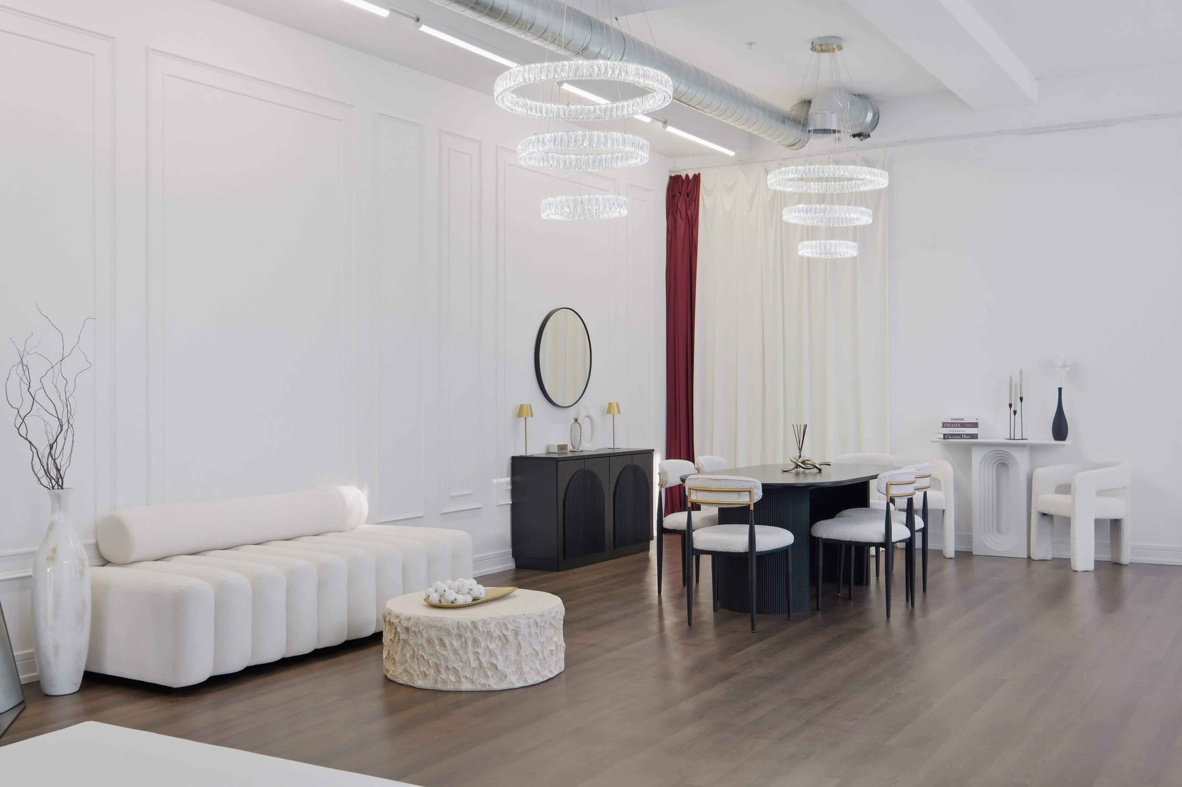 The image depicts a modern, minimalistic interior space featuring a white sofa, a round coffee table, a dining area with a black table and white chairs, and elegant light fixtures hanging from the ceiling.