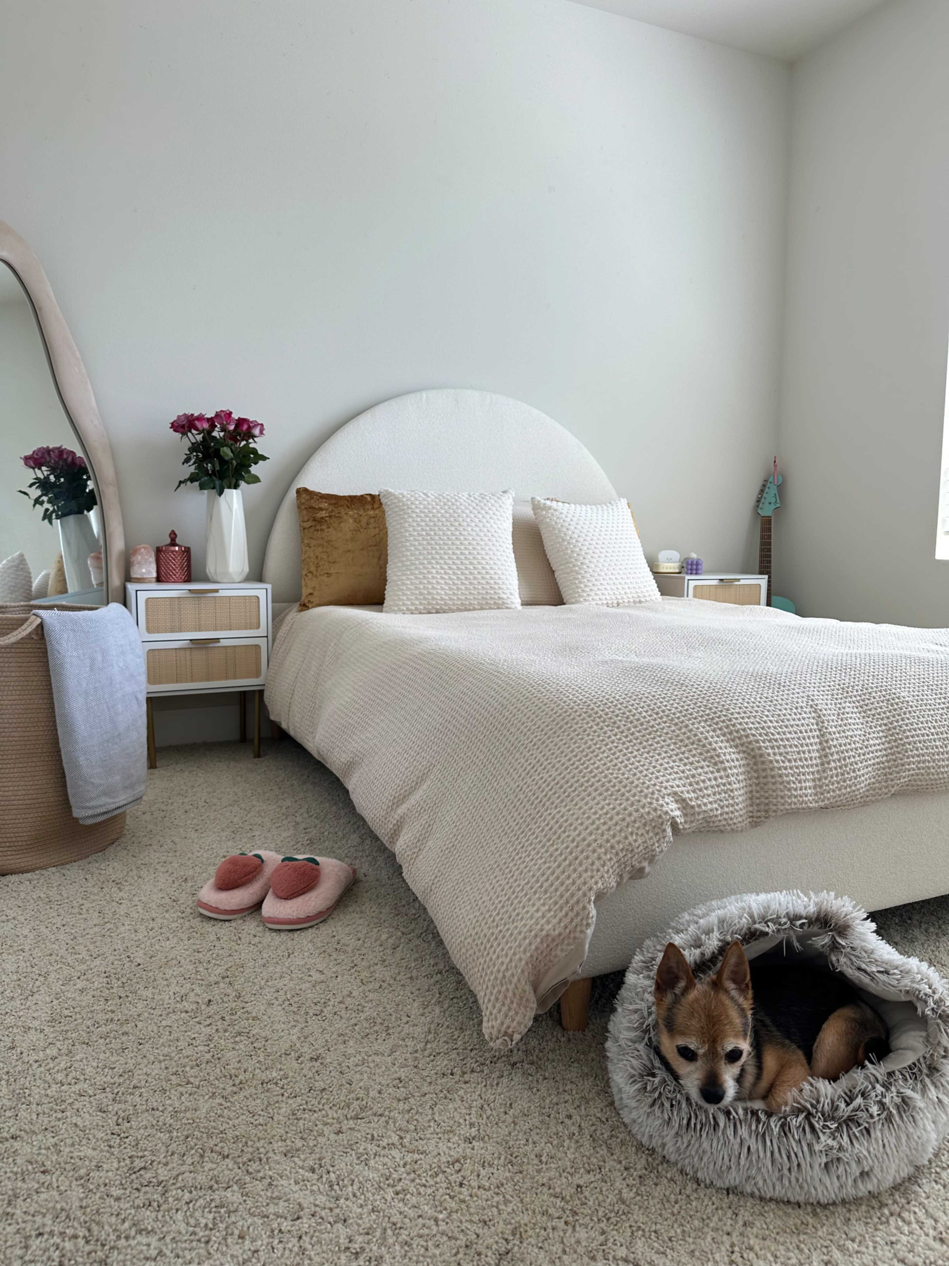 A neatly arranged bedroom features a bed with beige bedding, a small dog resting in a cozy bed, and bedside tables adorned with flowers and decor.