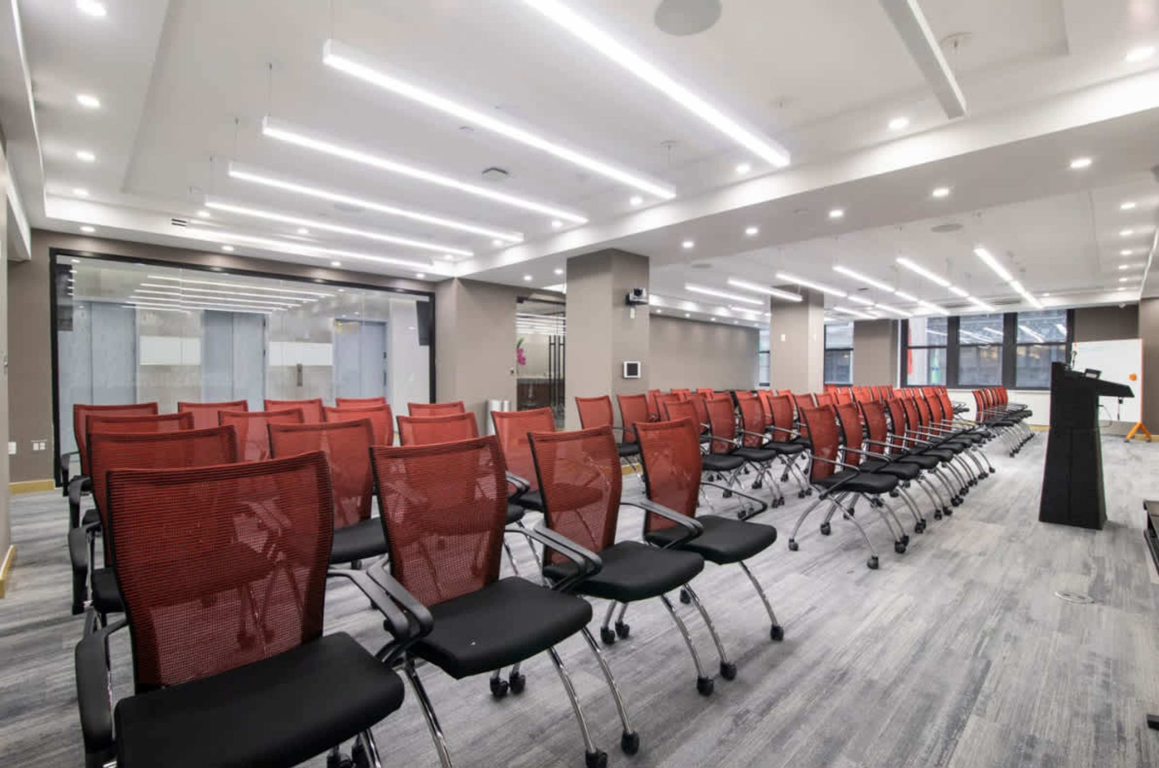 The image shows a conference room with rows of red mesh chairs set up in front of a podium and large windows.