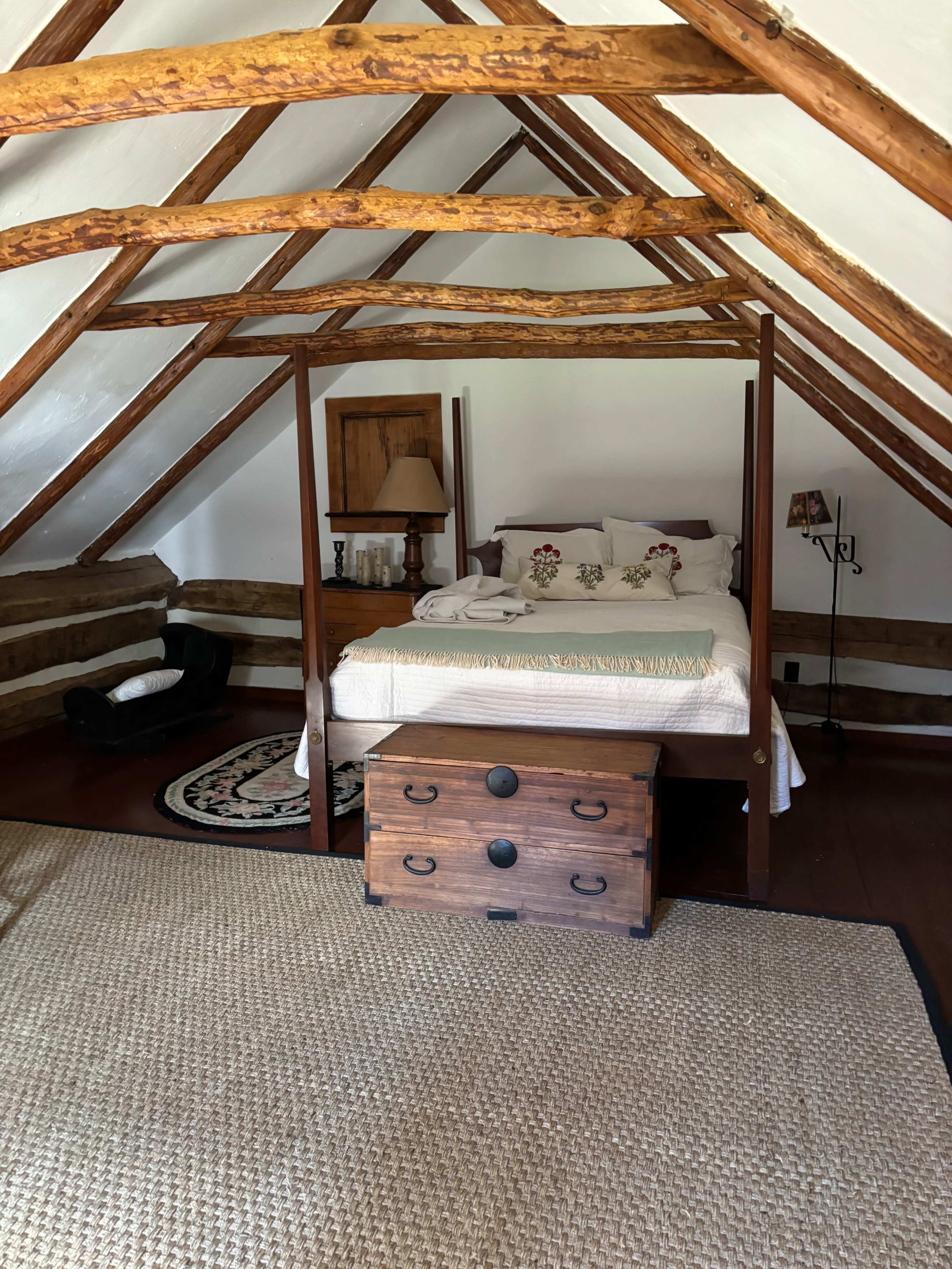 A cozy attic bedroom features a four-poster bed, wooden beams, and a vintage trunk at the foot of the bed.