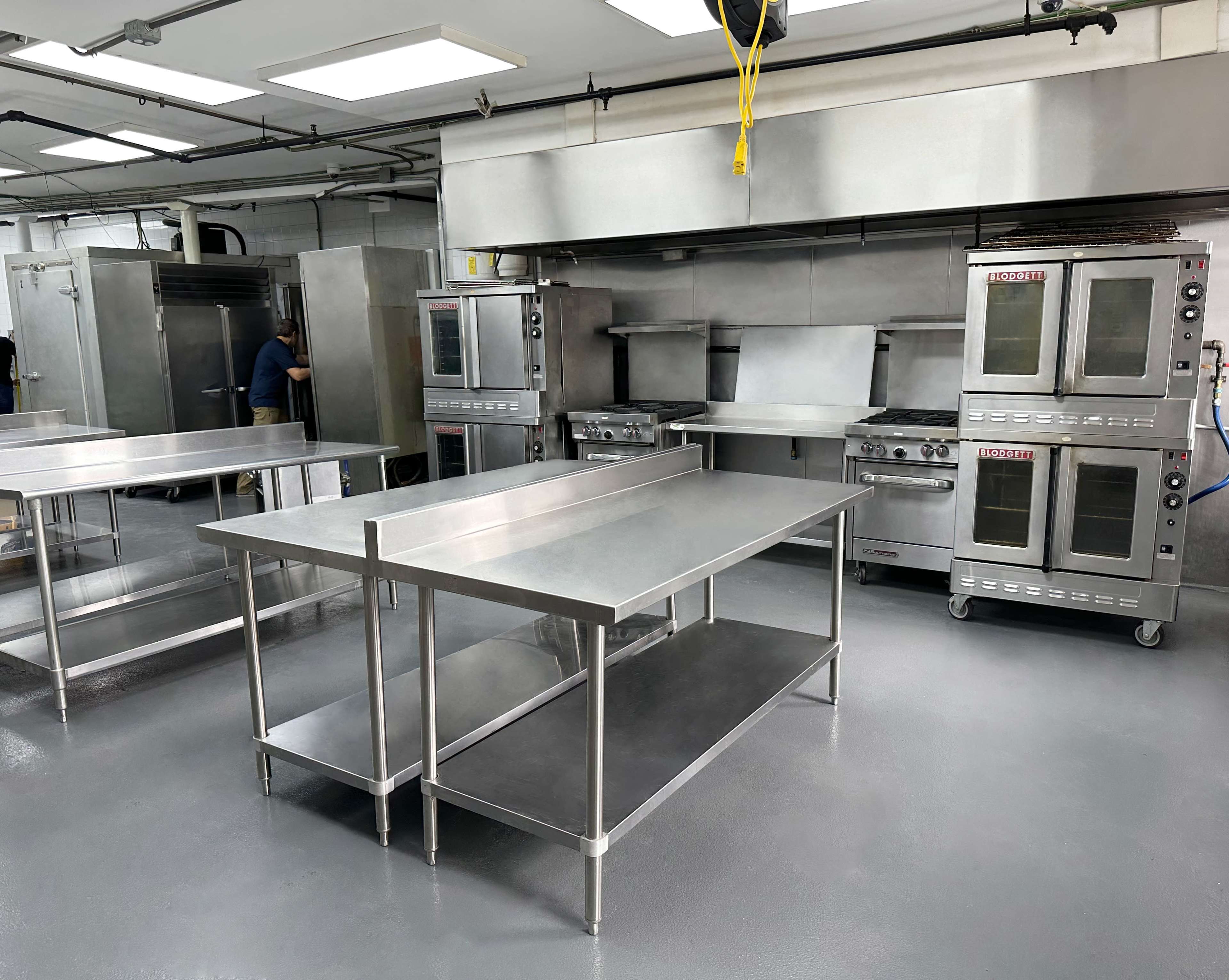 The image shows a commercial kitchen with stainless steel counters and appliances, including ovens and a cooktop, in a clean and organized layout.