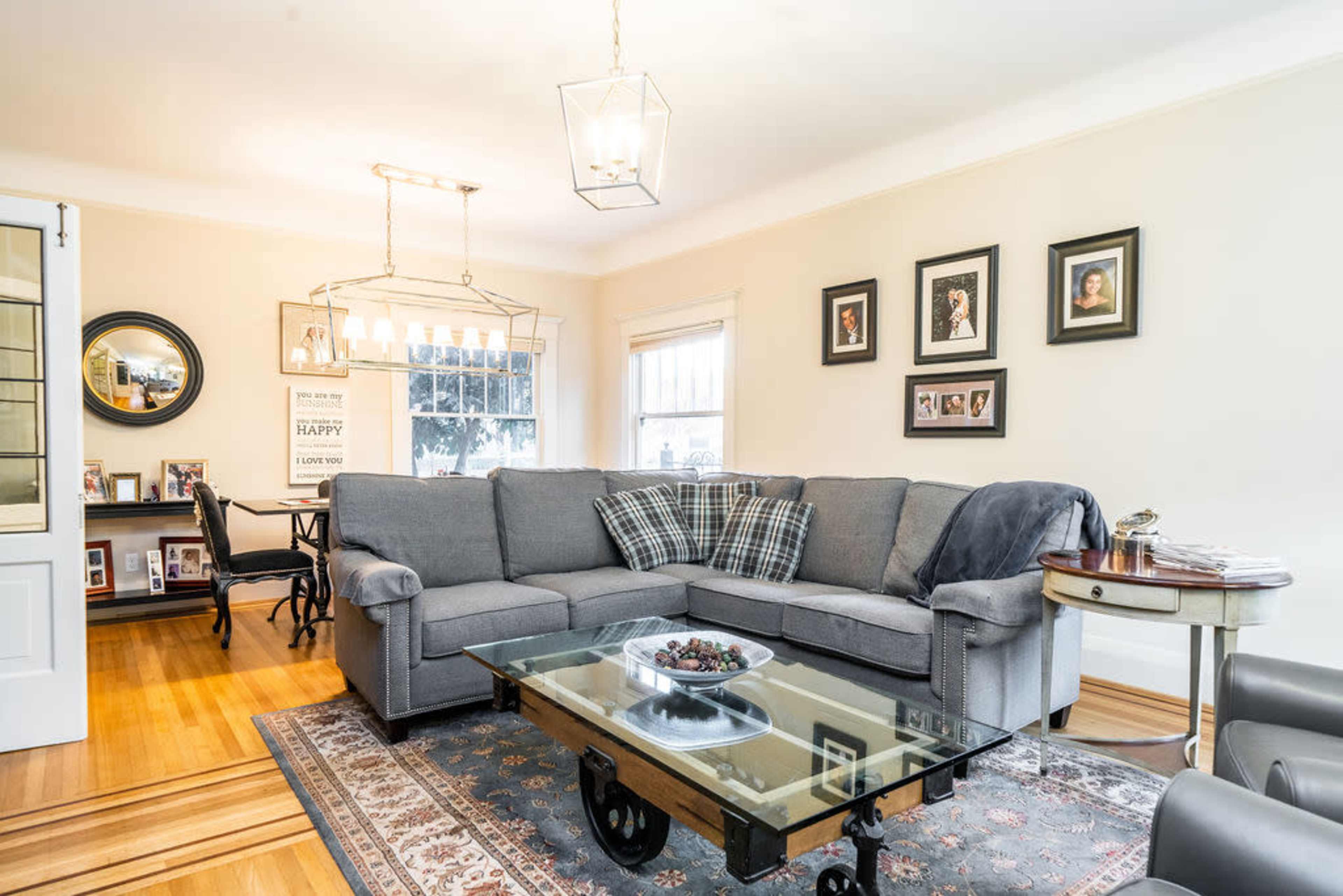The image shows a cozy living room featuring a gray sectional sofa, a glass coffee table, and framed photographs on the walls, with wooden flooring and a dining area in the background.