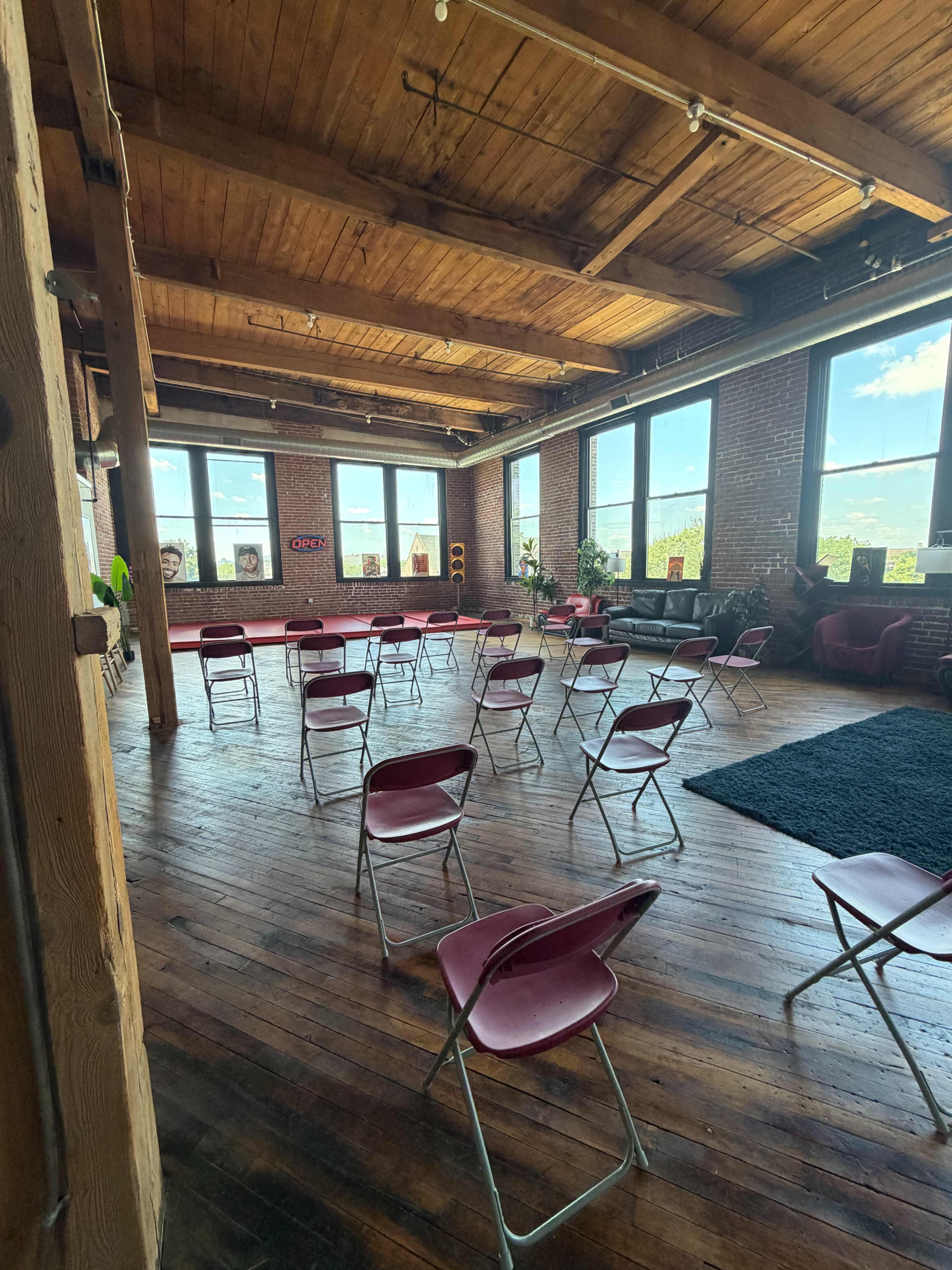 The image shows a spacious room with wooden floors, large windows, and several red folding chairs arranged in a circular pattern around a central area.
