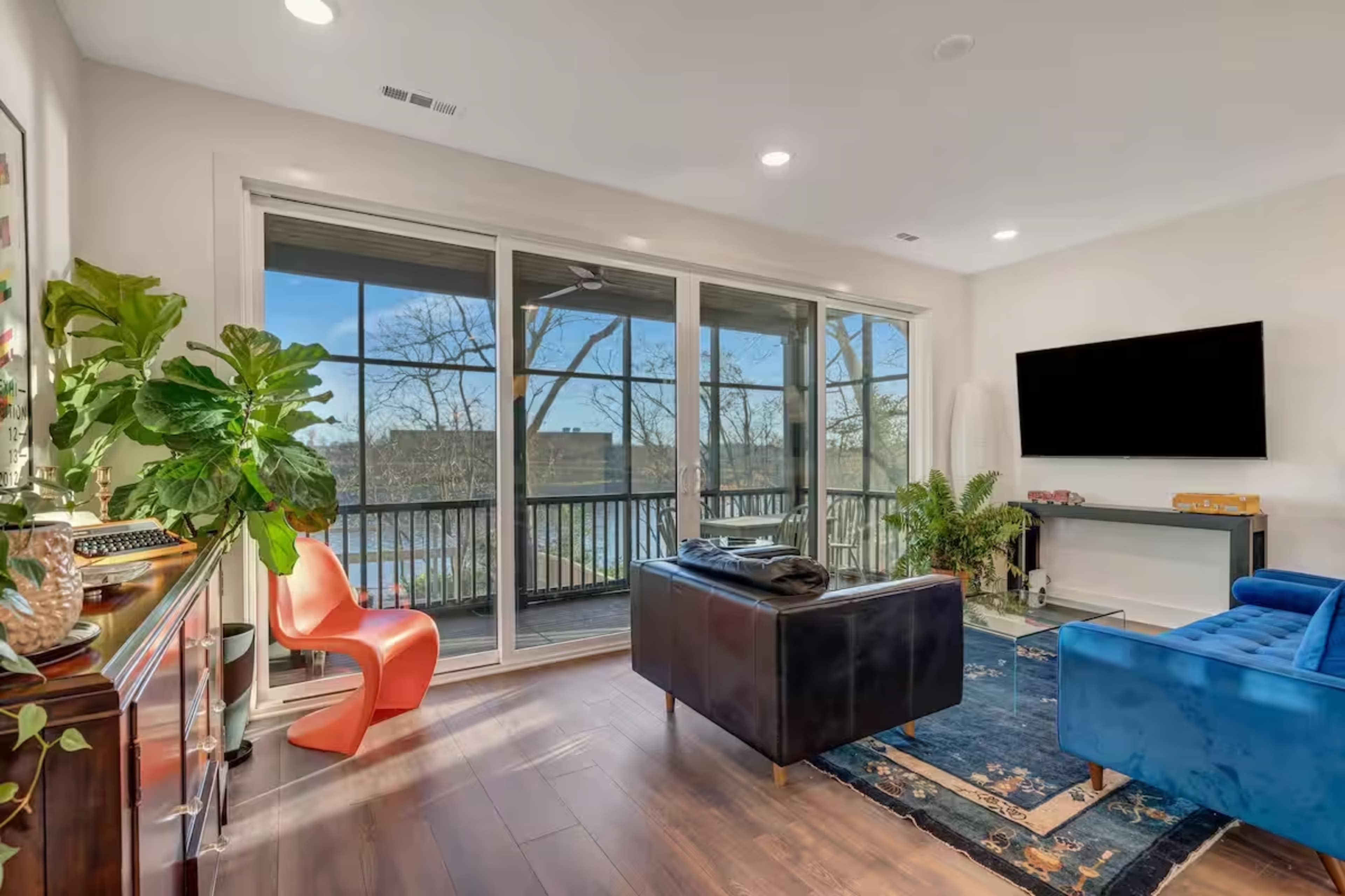 The image shows a modern living room with large windows overlooking a river, furnished with a blue couch, a black lounge chair, and a bright orange chair.