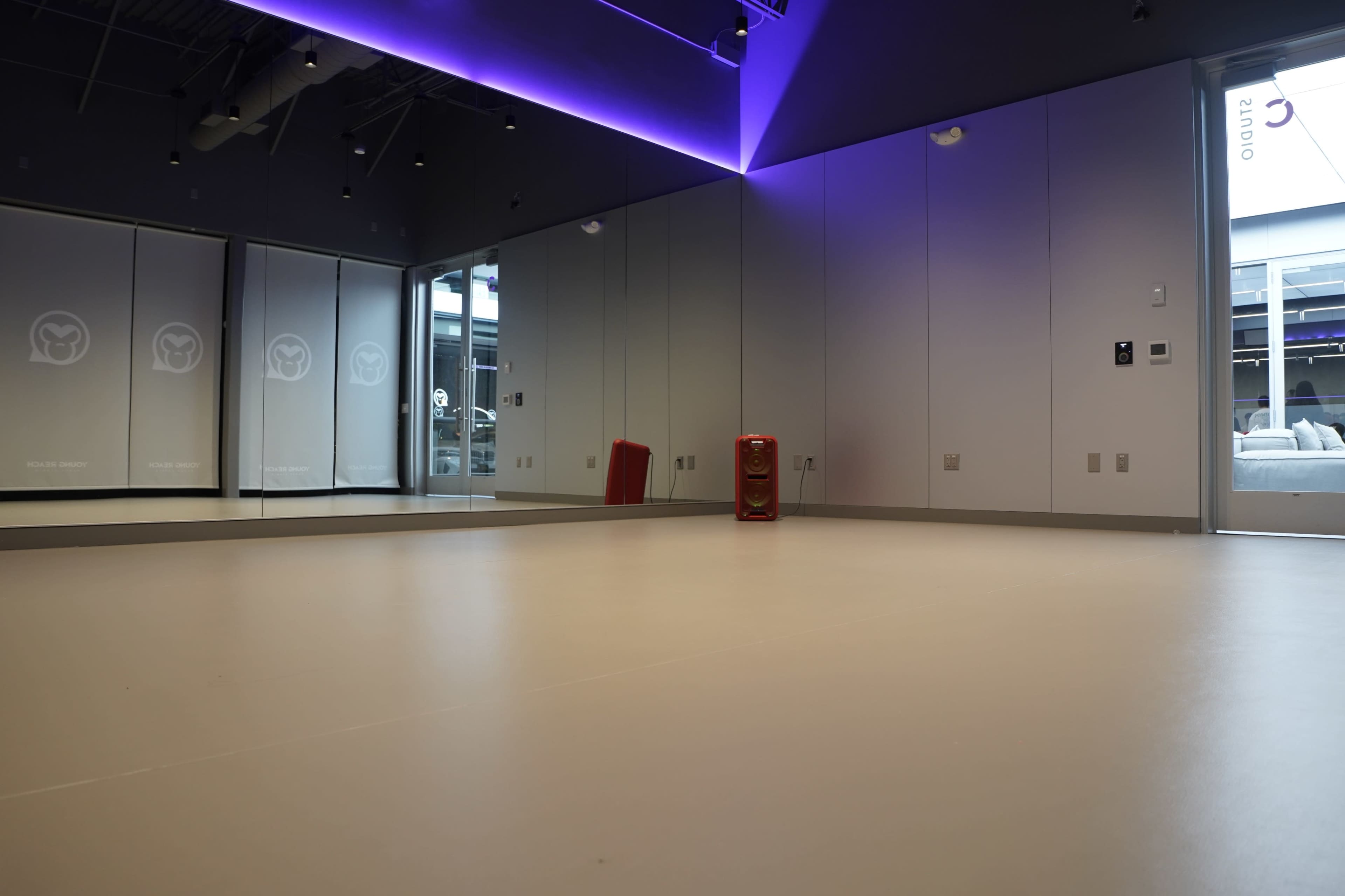 The image shows a spacious, minimally decorated studio with a large mirror, smooth flooring, and ambient lighting.