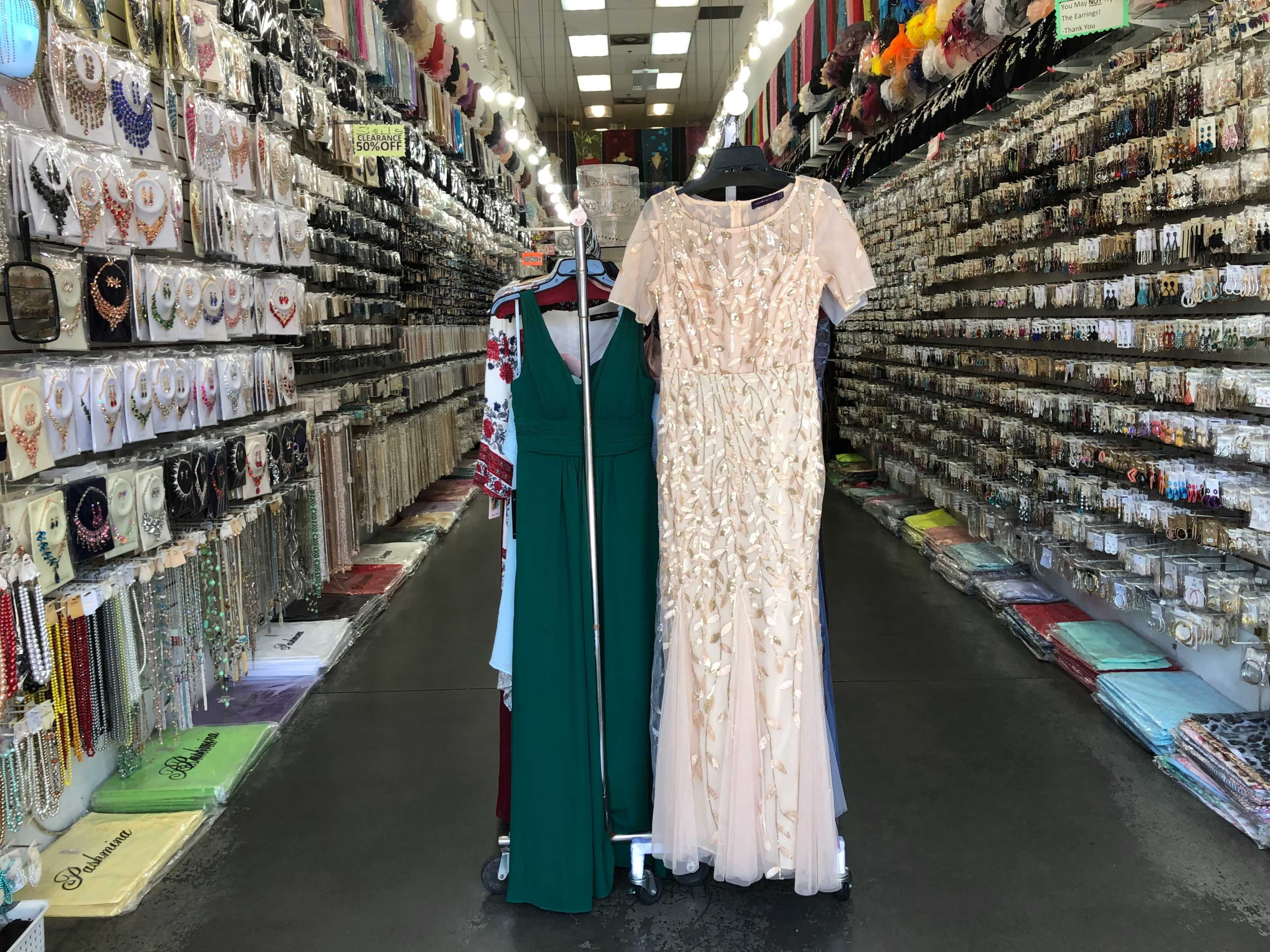 The image shows a narrow retail aisle lined with rows of colorful accessories and garments, featuring a rack displaying two formal dresses at the center.