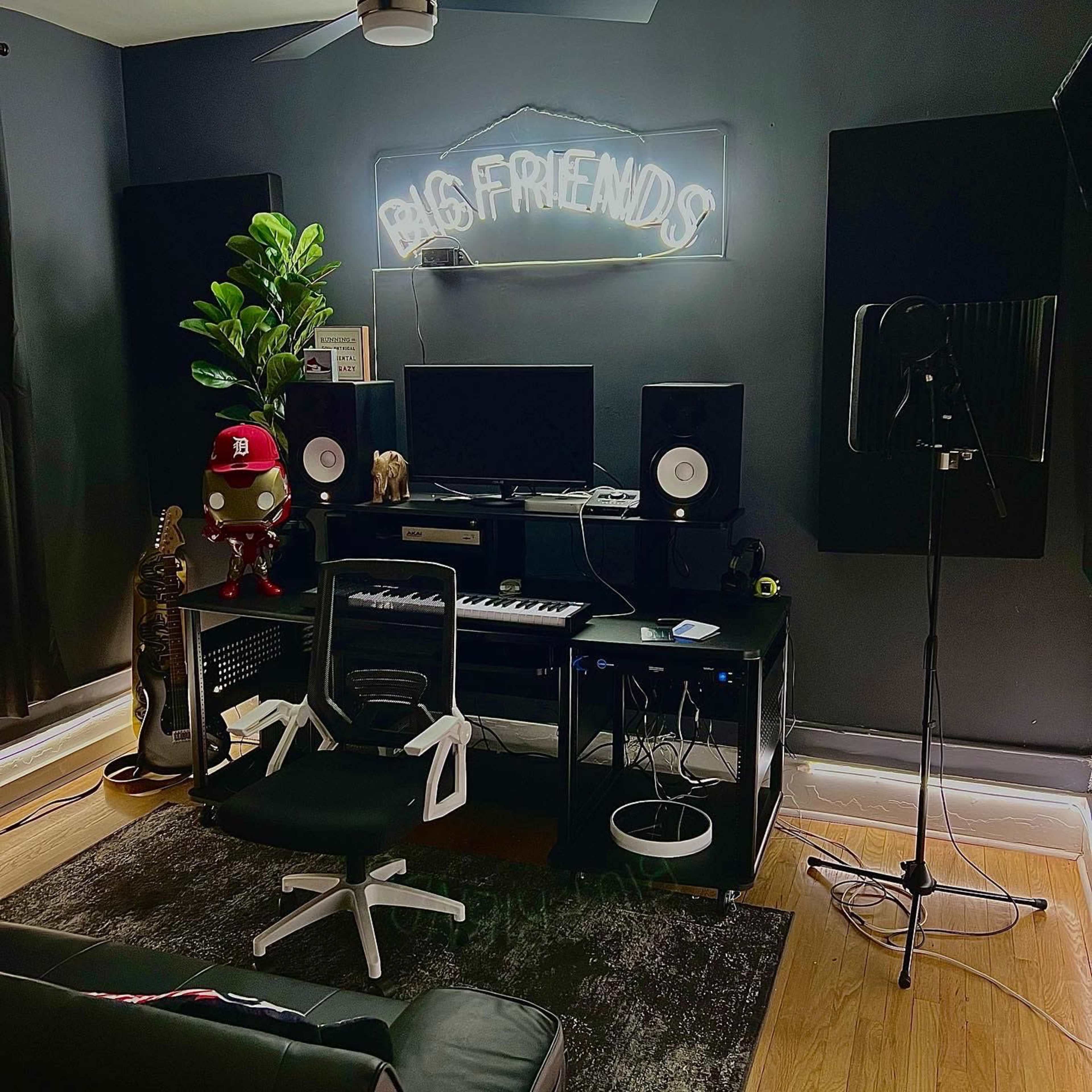A music studio featuring a desk with speakers, a computer, a microphone on a stand, and a guitar, all set against dark walls with a "BIG FRIENDS" neon sign.
