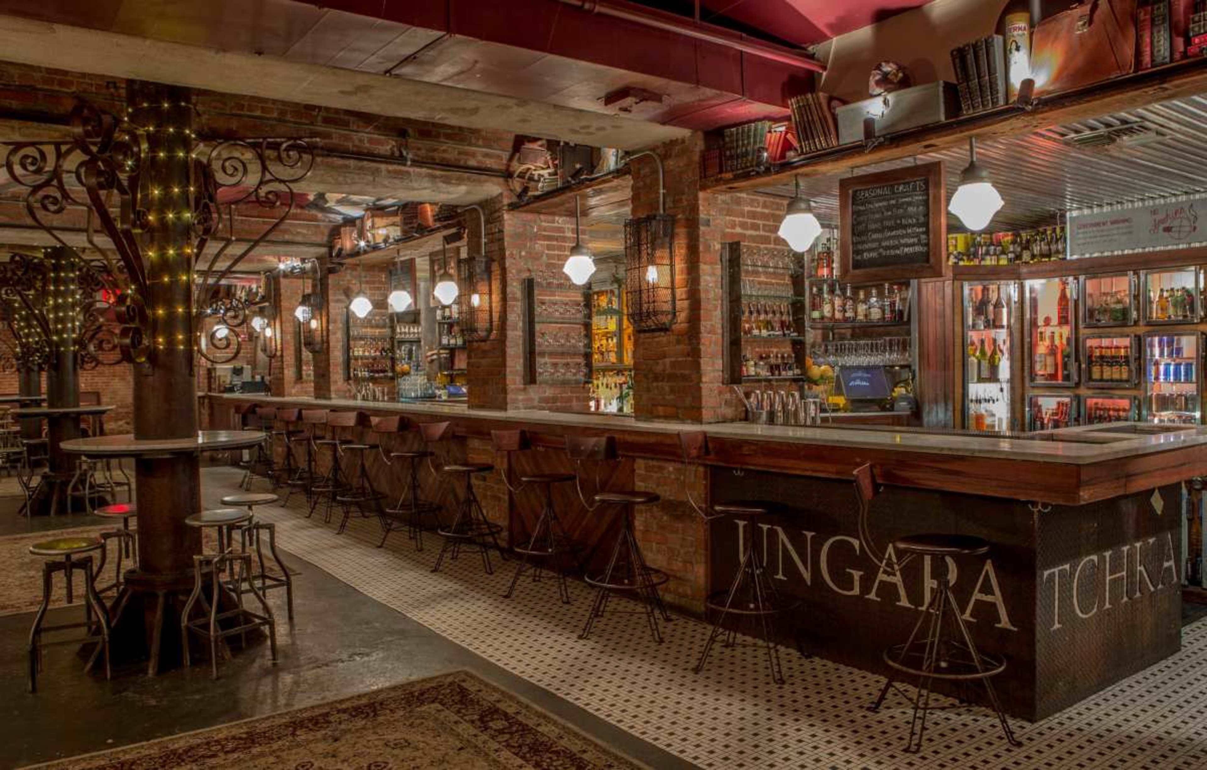 The image shows a stylish bar with a wooden counter and high stools, decorated with exposed brick walls and warm lighting.