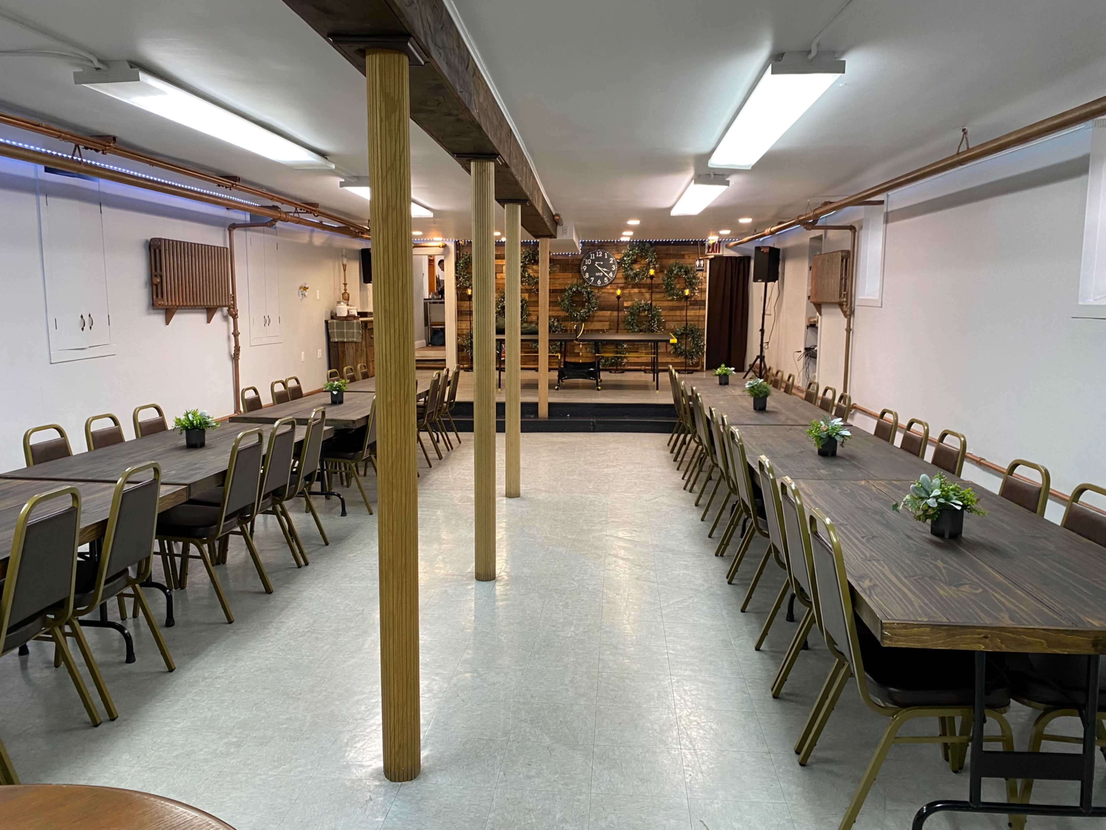 A large meeting room with long tables arranged in two parallel lines, surrounded by simple chairs and decorated with small plants on each table.