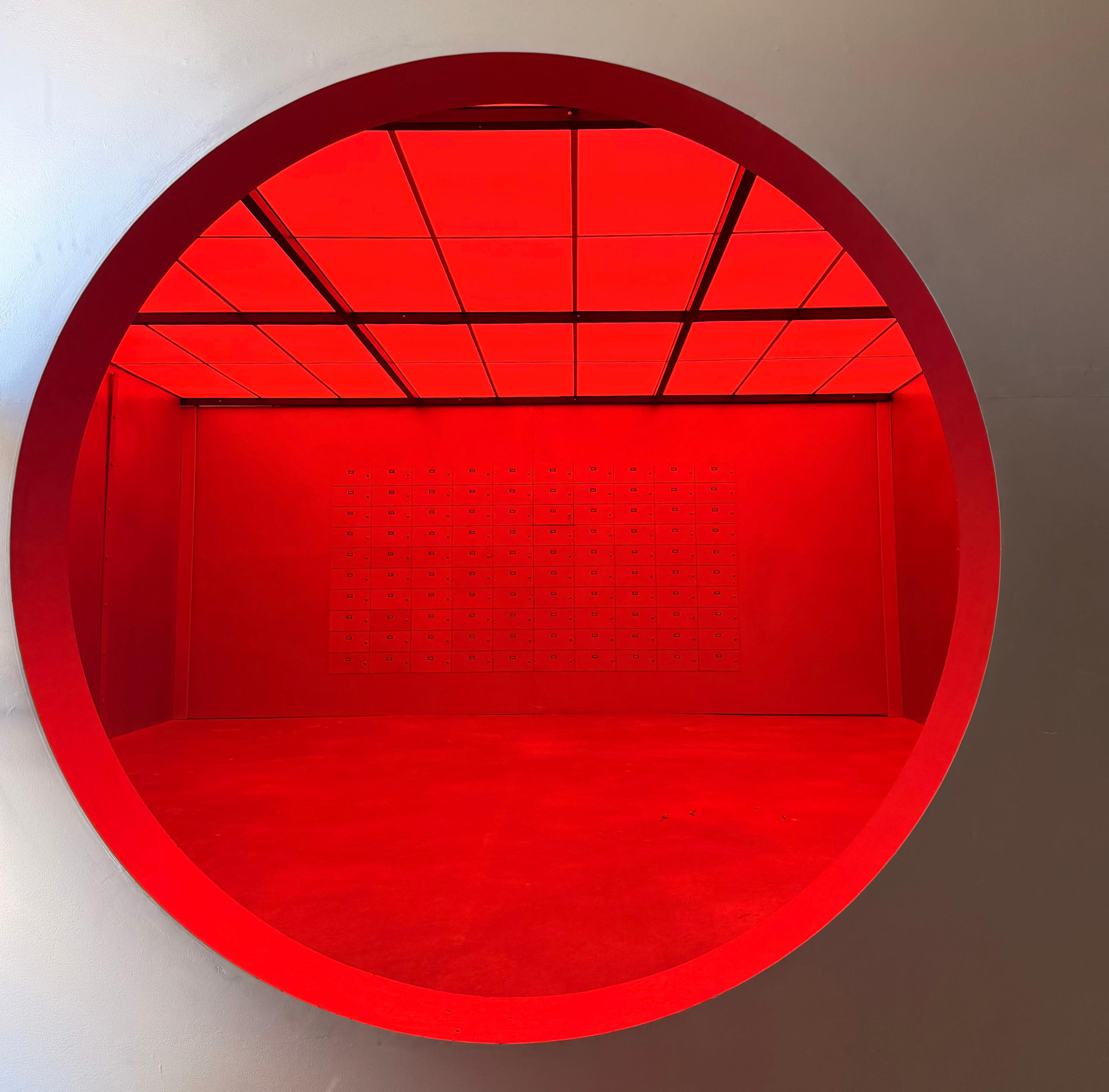 The image shows a circular opening in a wall that reveals a brightly lit red room with a grid of small boxes on the back wall.