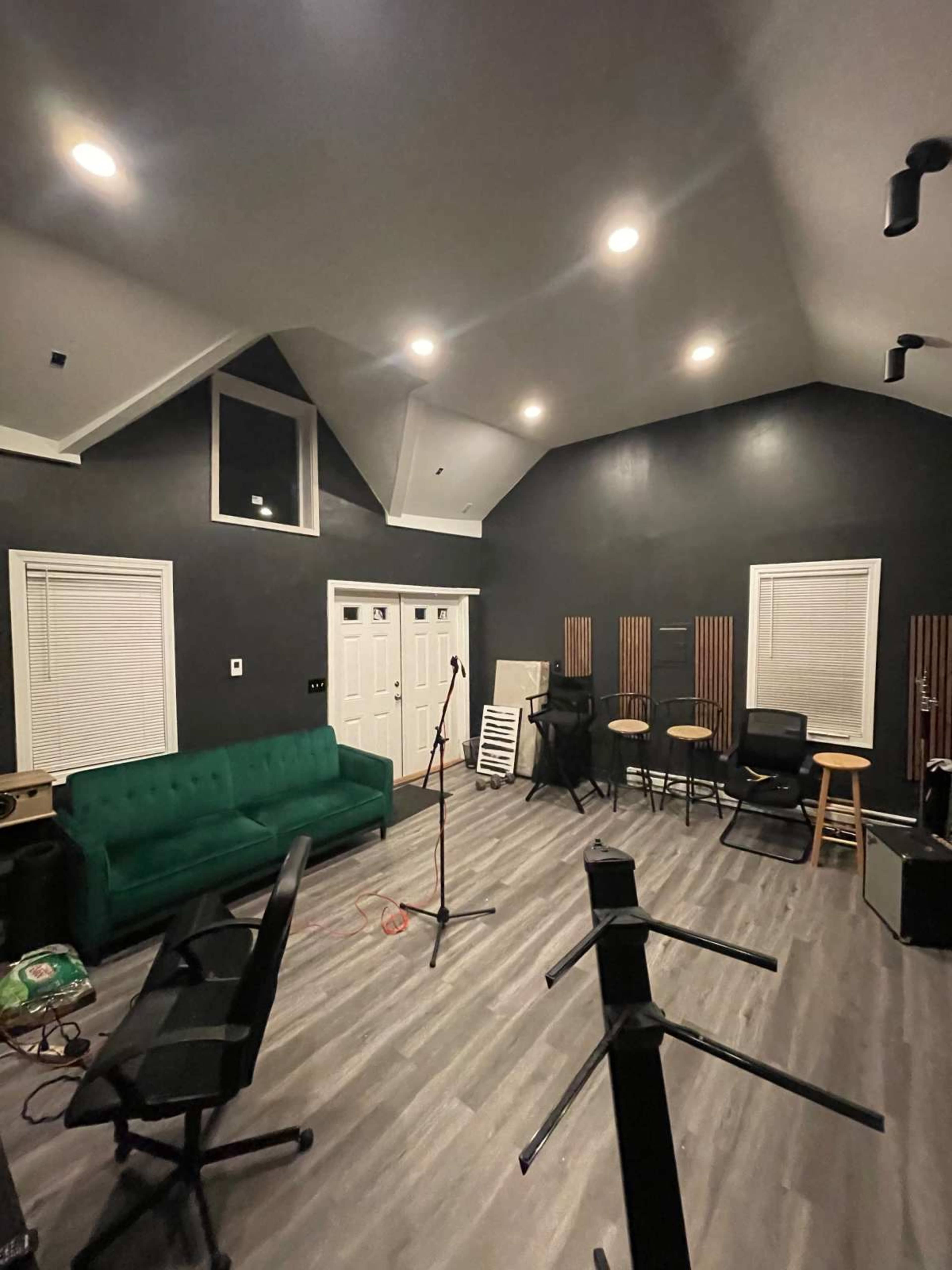 The image shows a dimly lit rehearsal space with a green couch, a microphone stand, several stools, and various musical equipment along the walls.