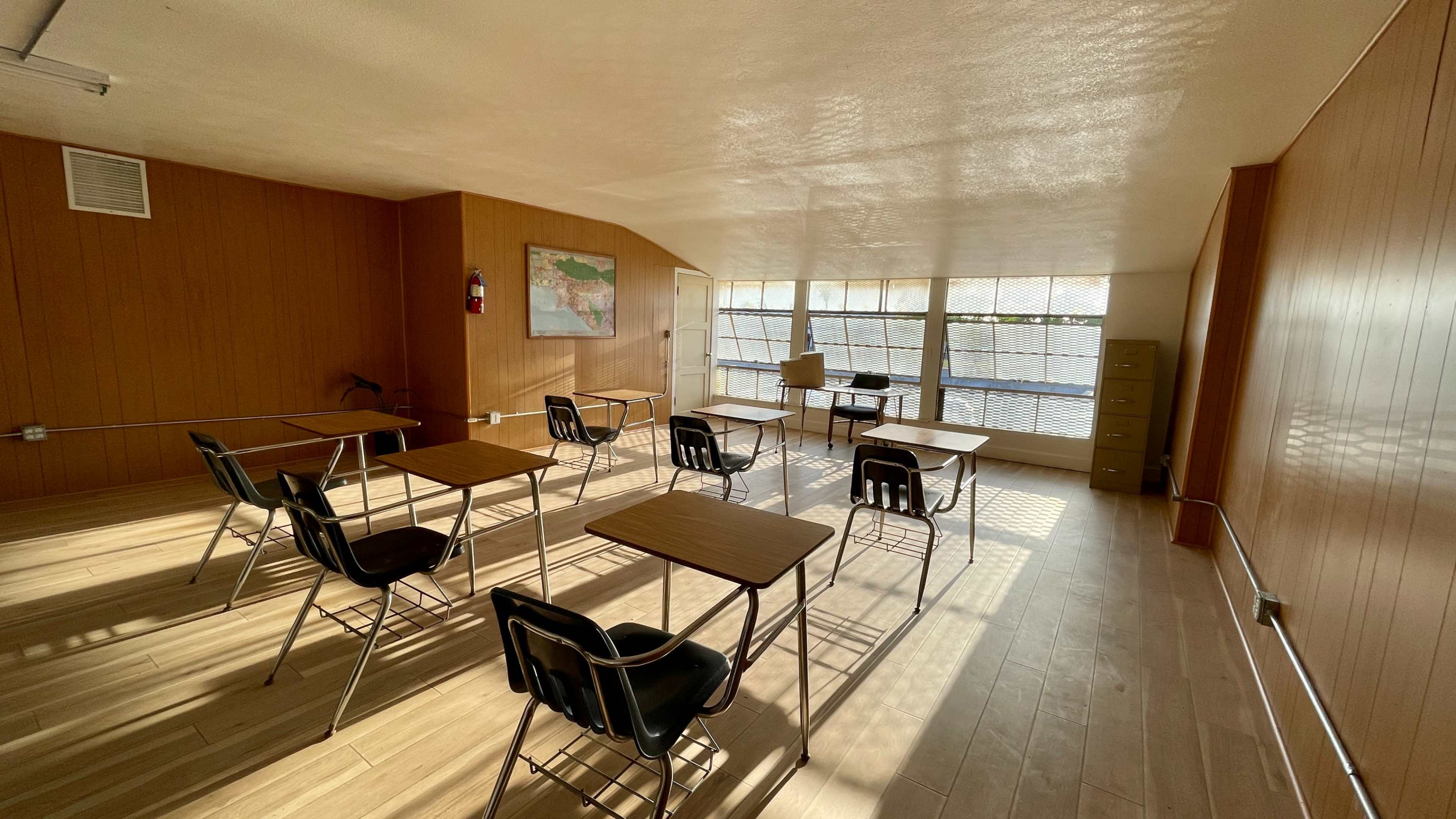 A classroom with several empty desks arranged around a central area, illuminated by natural light from large windows.