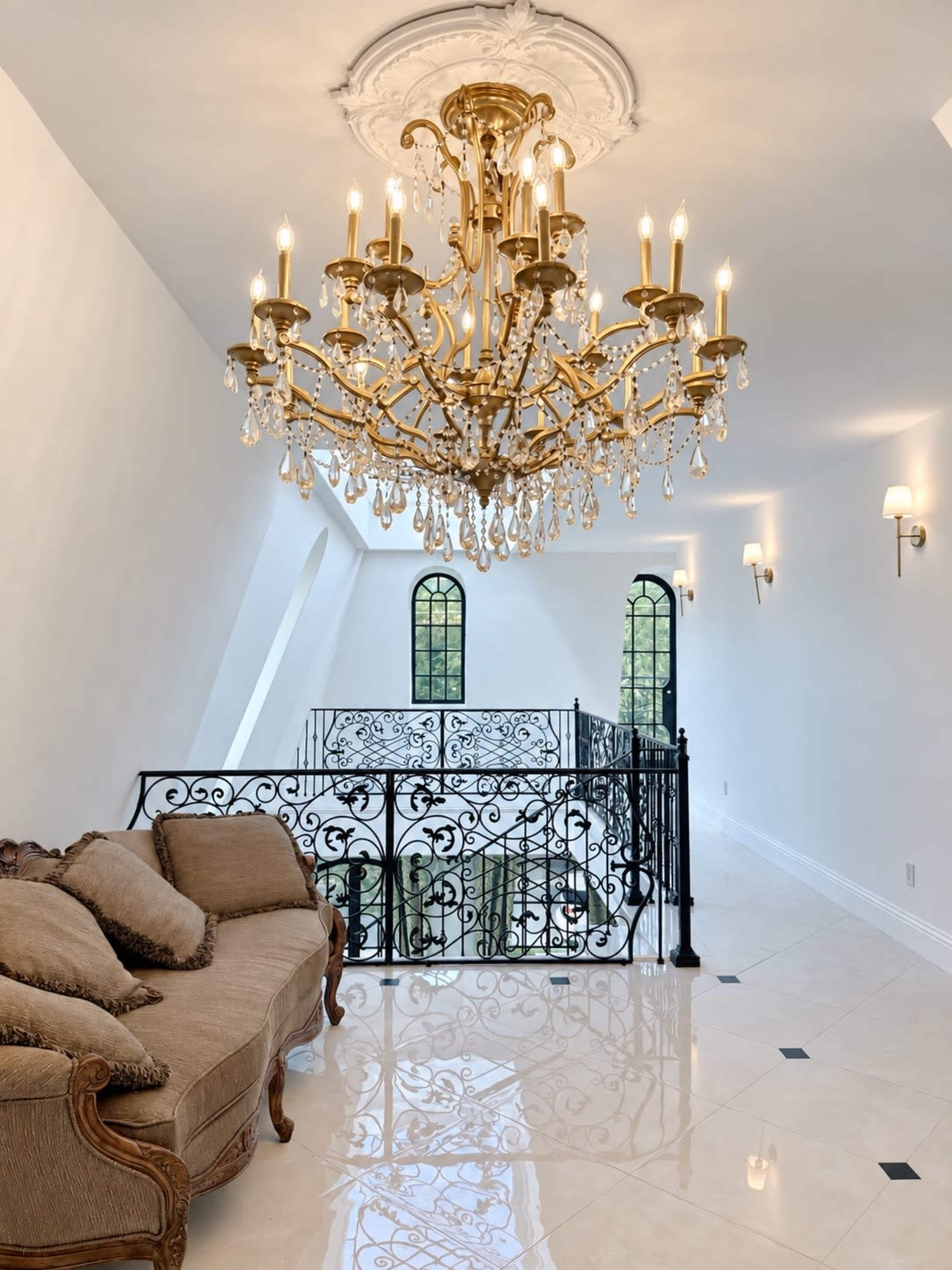 The image shows a spacious interior with a decorative chandelier hanging from a ceiling, a brown couch on one side, and an ornate black railing overlooking a lower level.