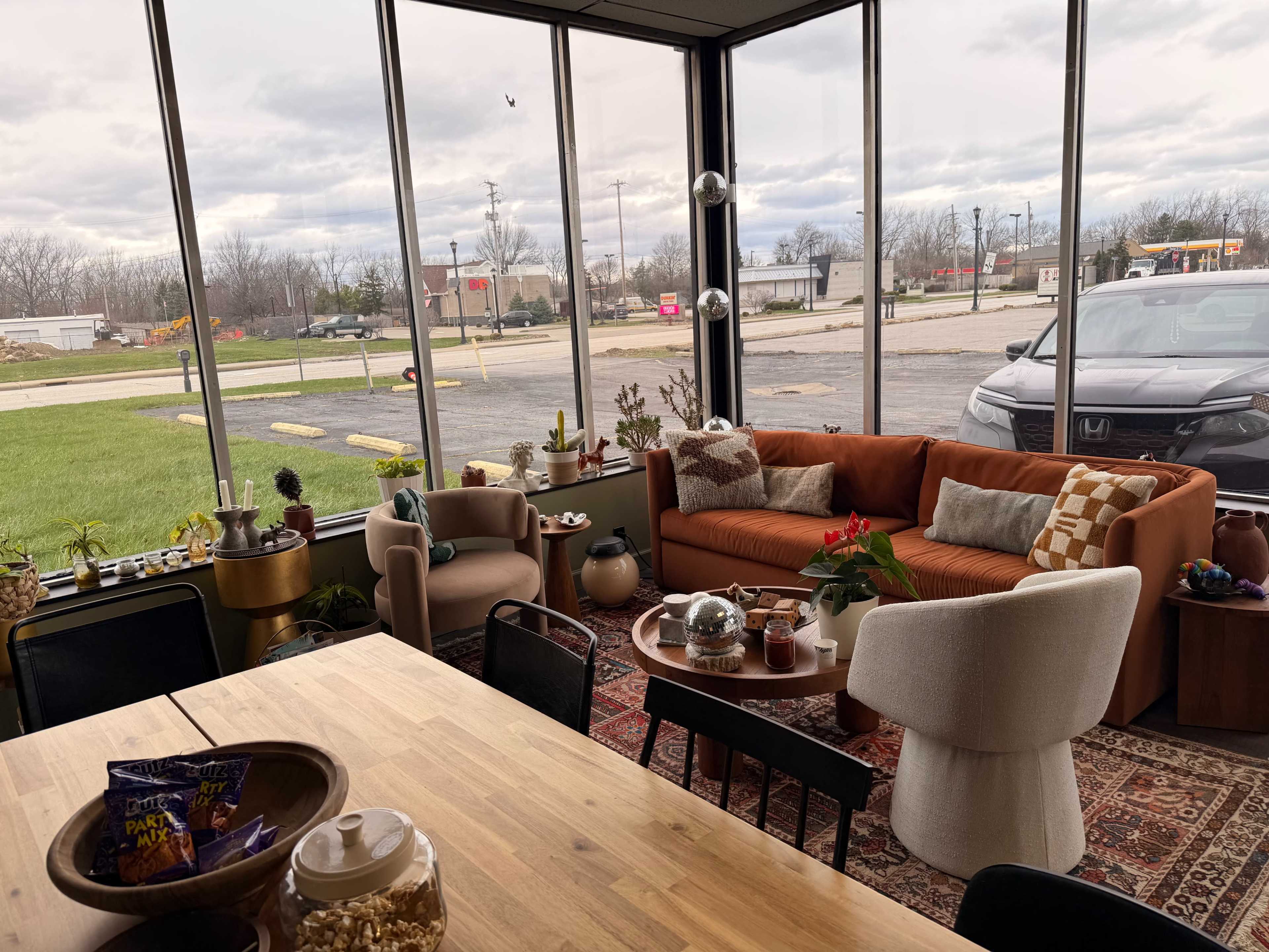 The image shows a cozy interior space with large windows, featuring a mix of modern furniture, including a plush orange sofa and various plants, alongside a dining table and snacks.