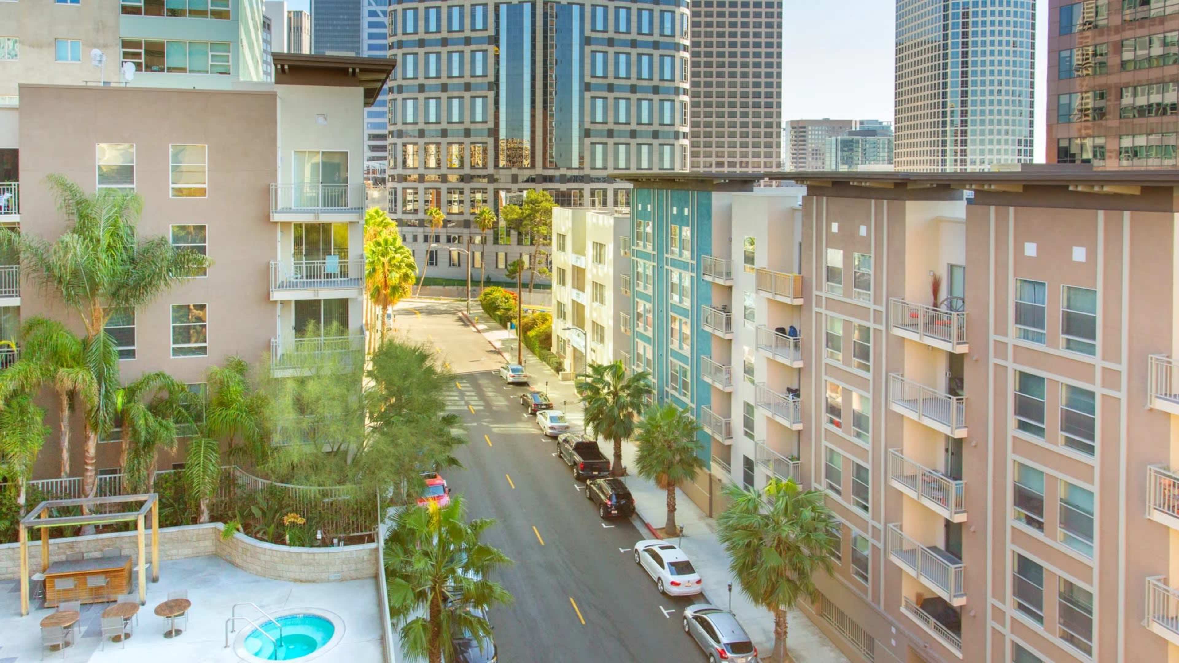The image shows a city street lined with modern apartment buildings and palm trees, leading toward a backdrop of high-rise buildings.