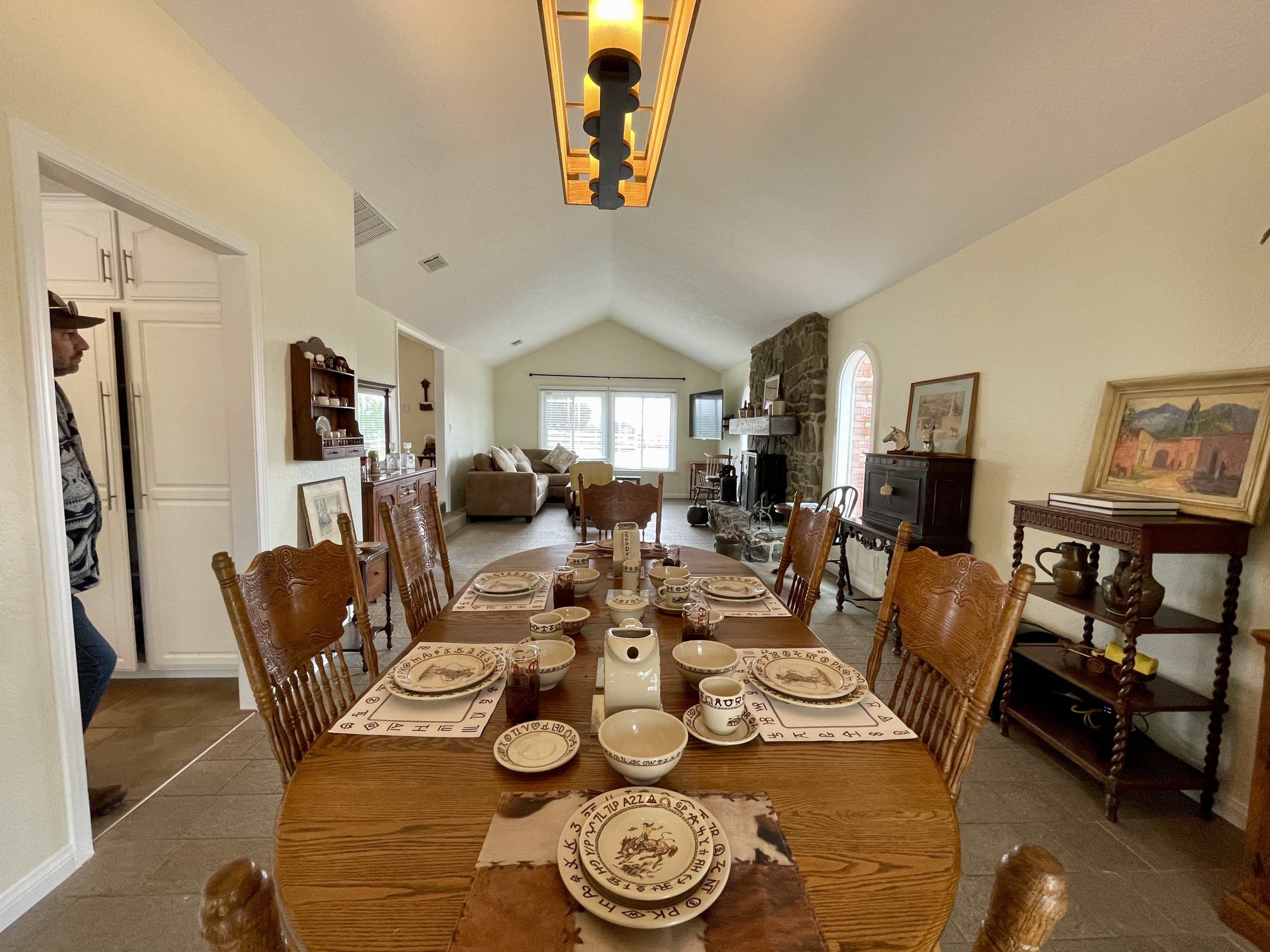 A dining table set for a meal with dishes and utensils is positioned in a spacious, open-concept living area featuring a stone fireplace and large windows.