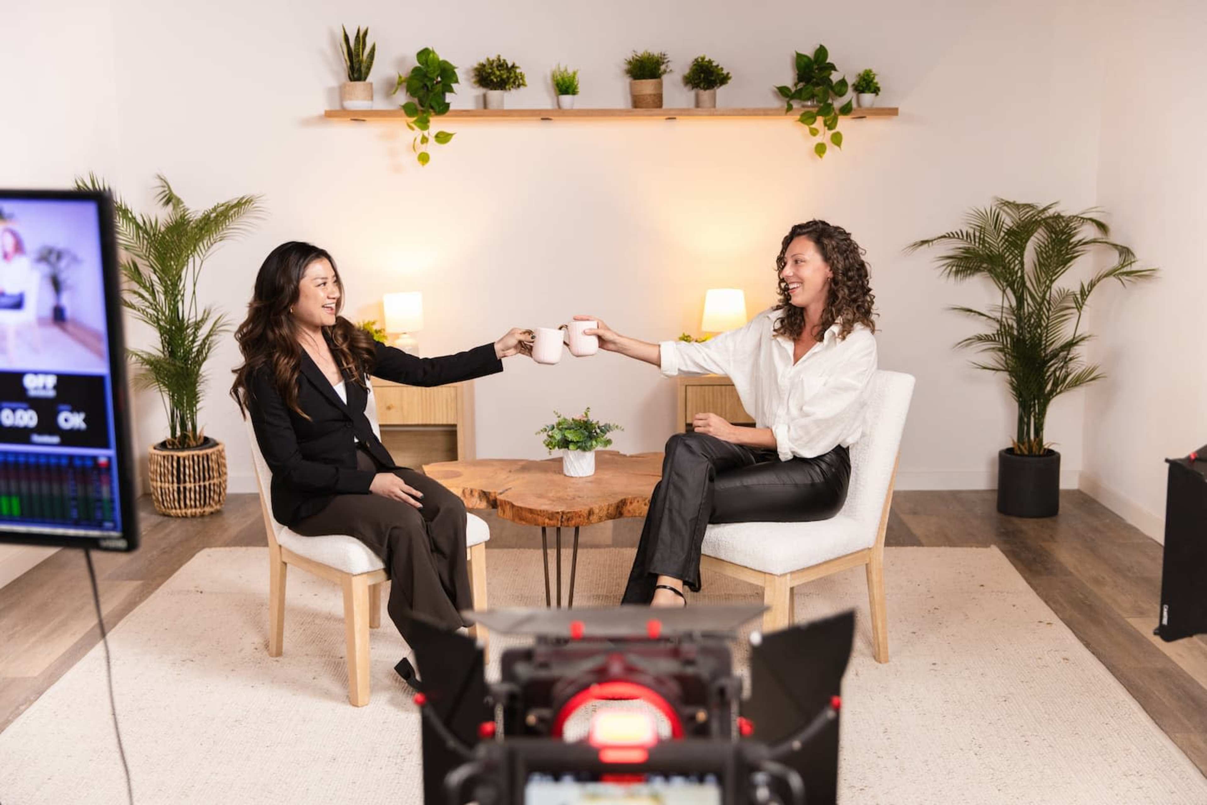Two women are seated in a well-lit studio, raising coffee mugs in a toast while sitting across from each other on chairs next to a small round table and surrounded by potted plants.