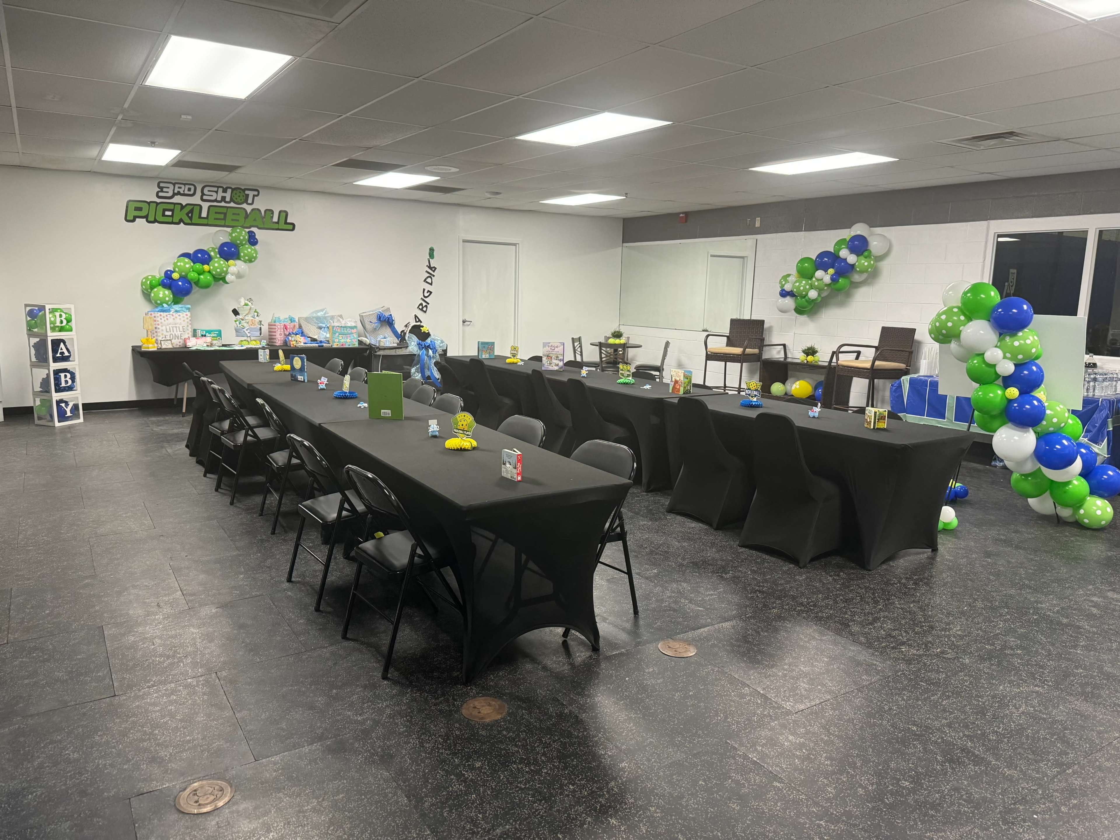 A spacious event room features multiple black tablecloth-covered tables arranged in two rows, adorned with balloons in blue, green, and white, and decorated with various centerpieces.