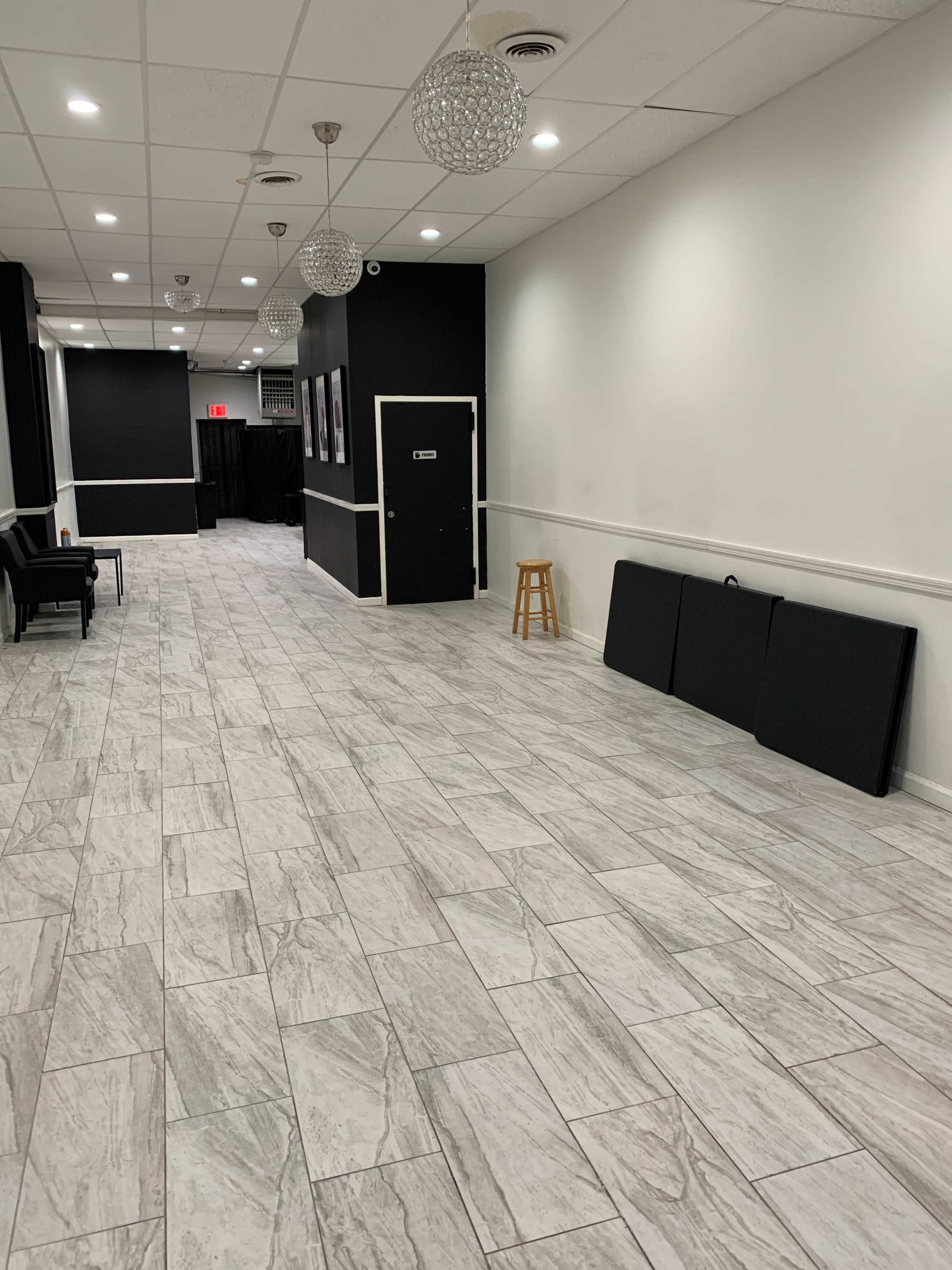 A long, empty hallway with tiled flooring, a few chairs on one side, and a stool near a closed door, with decorative light fixtures overhead.